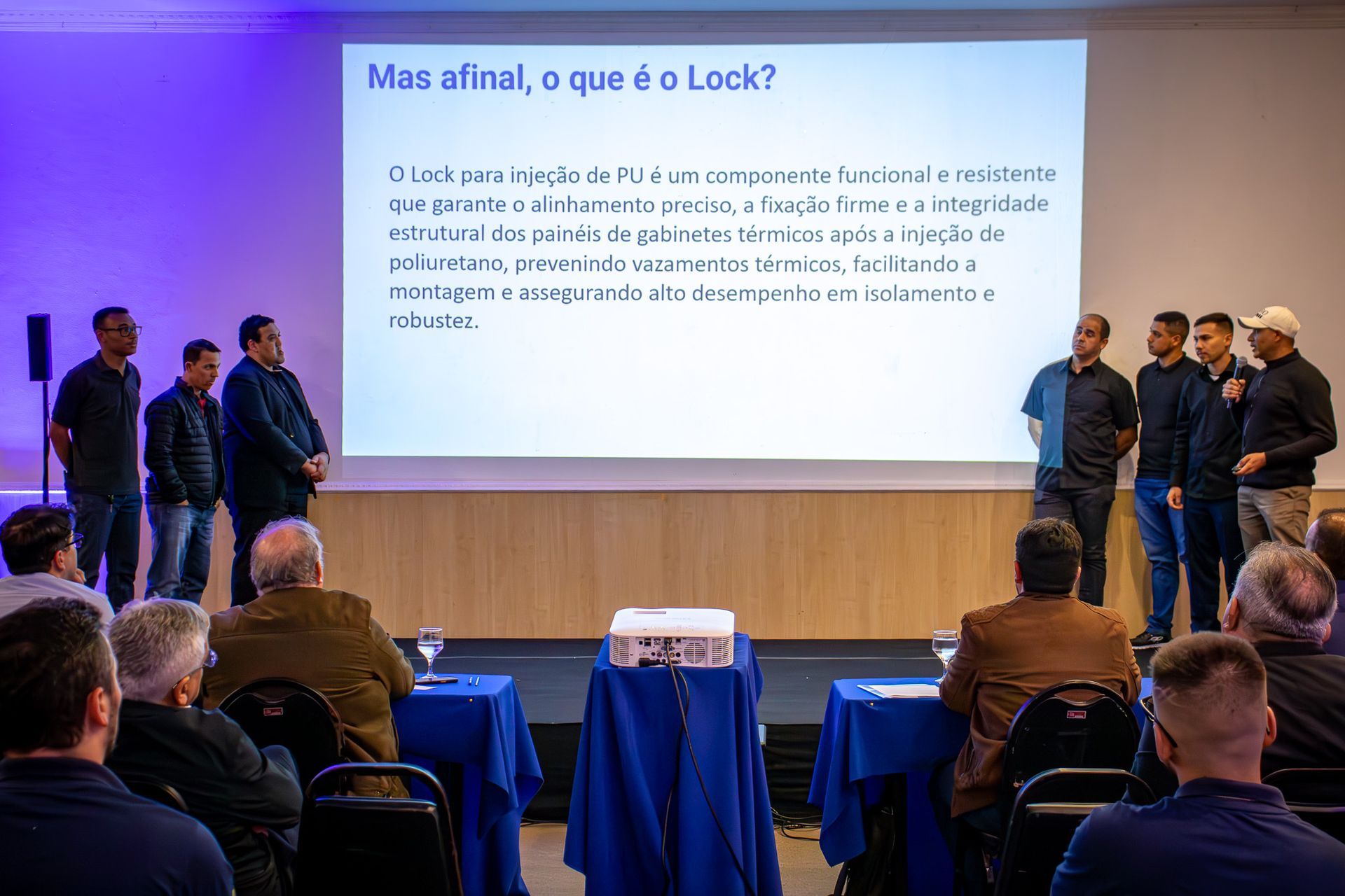 Uma apresentação com pessoas em frente a uma tela exibindo texto. Um grupo está no palco, de frente para o público sentado em mesas.