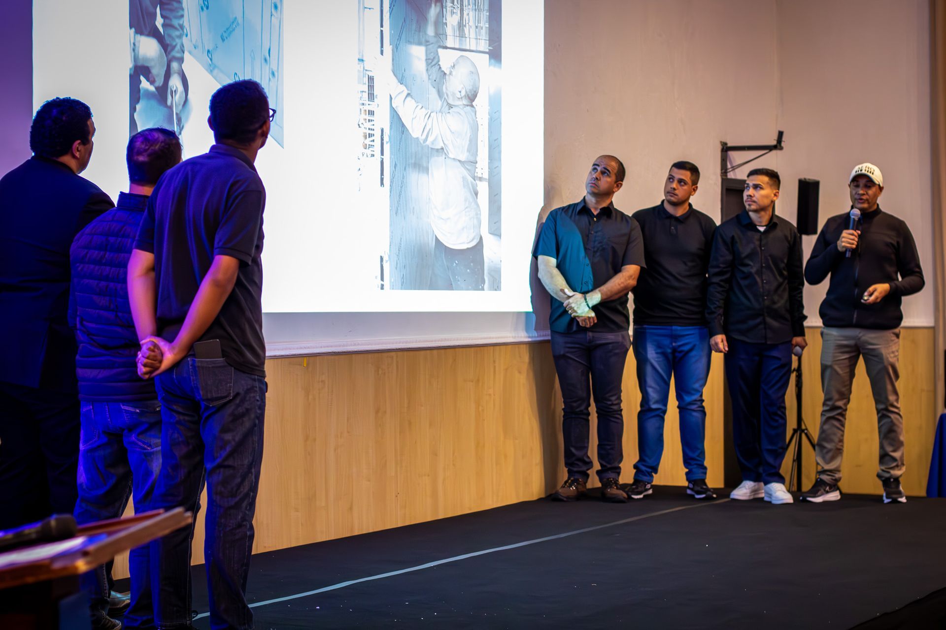 Grupo de homens se apresentando diante de uma tela com uma imagem projetada. Alguns homens estão em pé no palco e outros na plateia.