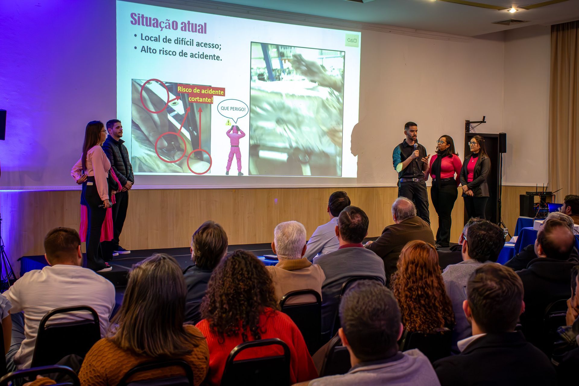 Uma apresentação com quatro pessoas no palco falando para uma plateia. A tela exibe um texto e a imagem de uma pessoa vestida de rosa.