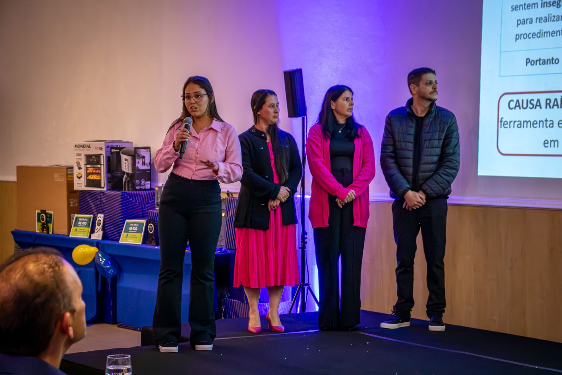 Quatro pessoas em um palco fazendo uma apresentação. Uma mulher fala ao microfone, enquanto outras pessoas ficam ao lado dela.