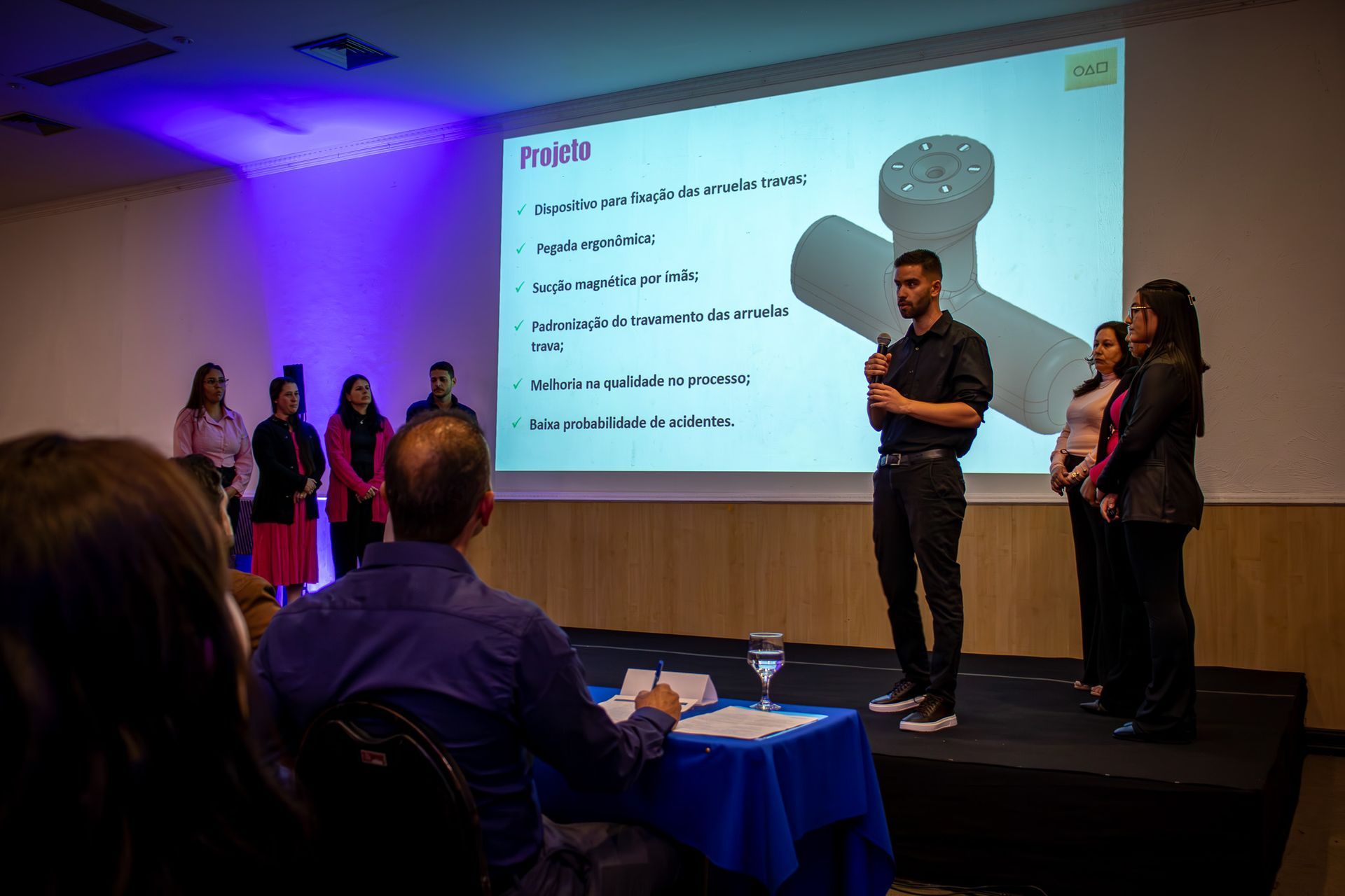 Um homem faz uma apresentação no palco com outras pessoas, enquanto um projetor exibe um projeto. O público assiste em uma sala iluminada com luzes azuis e brancas.