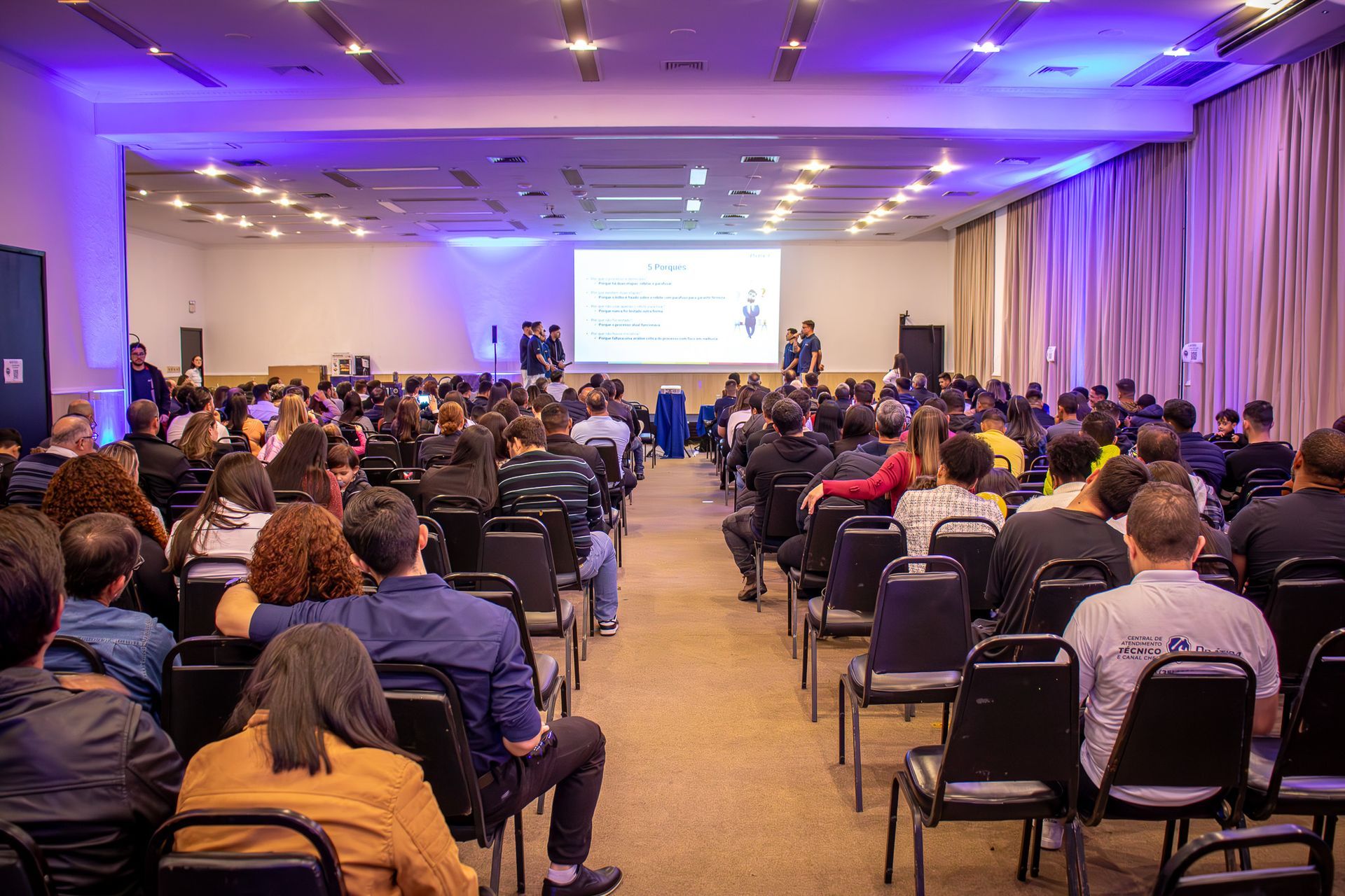 Uma grande sala de conferências com uma plateia de frente para uma tela de projeção. Os apresentadores estão em pé no palco, banhados por uma iluminação roxa.