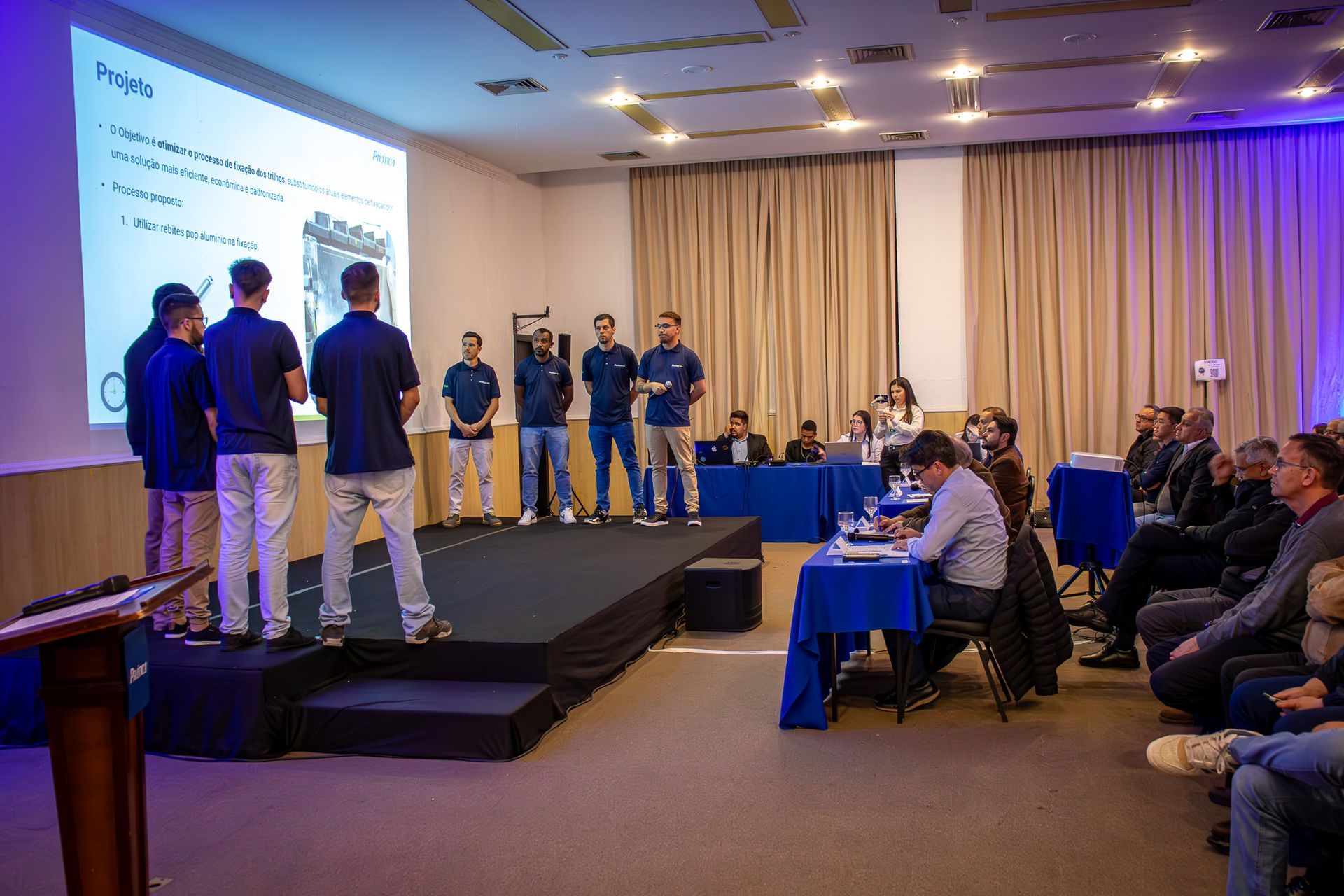 Uma apresentação é feita por um grupo de homens em um palco para uma plateia em uma sala de conferências.