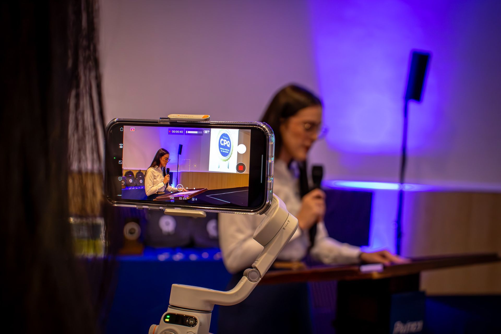 Uma mulher fazendo uma apresentação, vista através da tela de um celular em um gimbal. O cenário é um evento com iluminação azul.