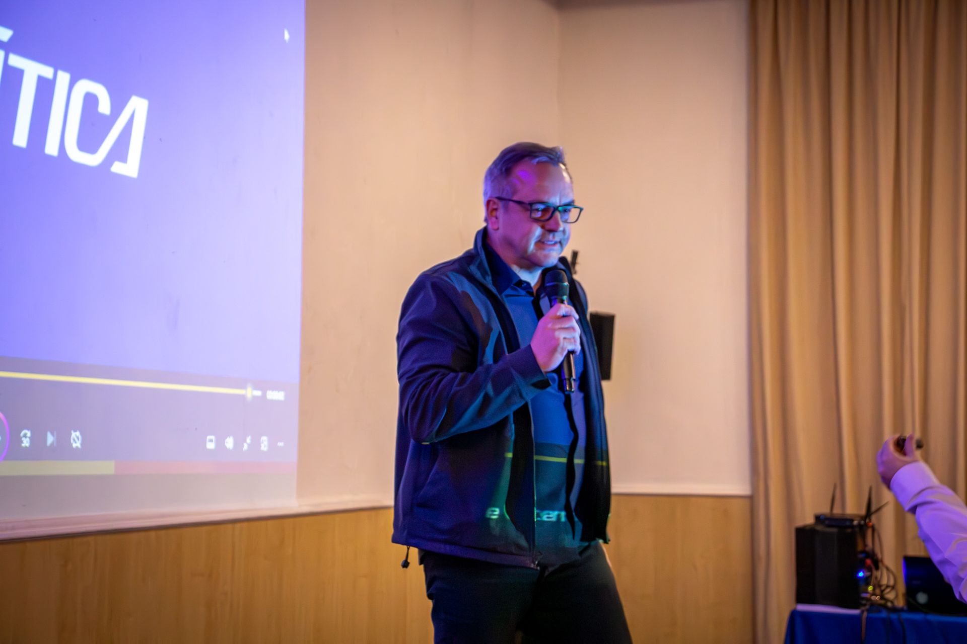 Homem de óculos falando em um microfone em uma sala de aula, em frente a uma tela exibindo texto.