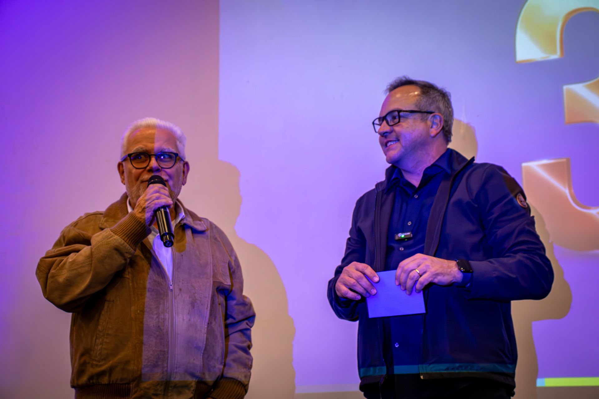 Dois homens no palco de um evento. Um, mais velho, fala ao microfone, vestindo uma jaqueta marrom. O outro segura um bilhete e sorri.