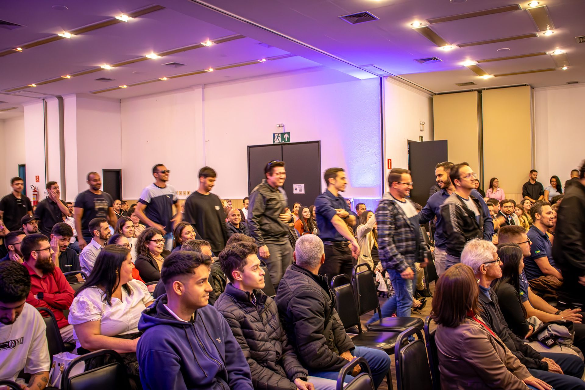 Um auditório lotado, cheio de pessoas assistindo a algo na frente. Iluminação fraca, pessoas sentadas e em pé.
