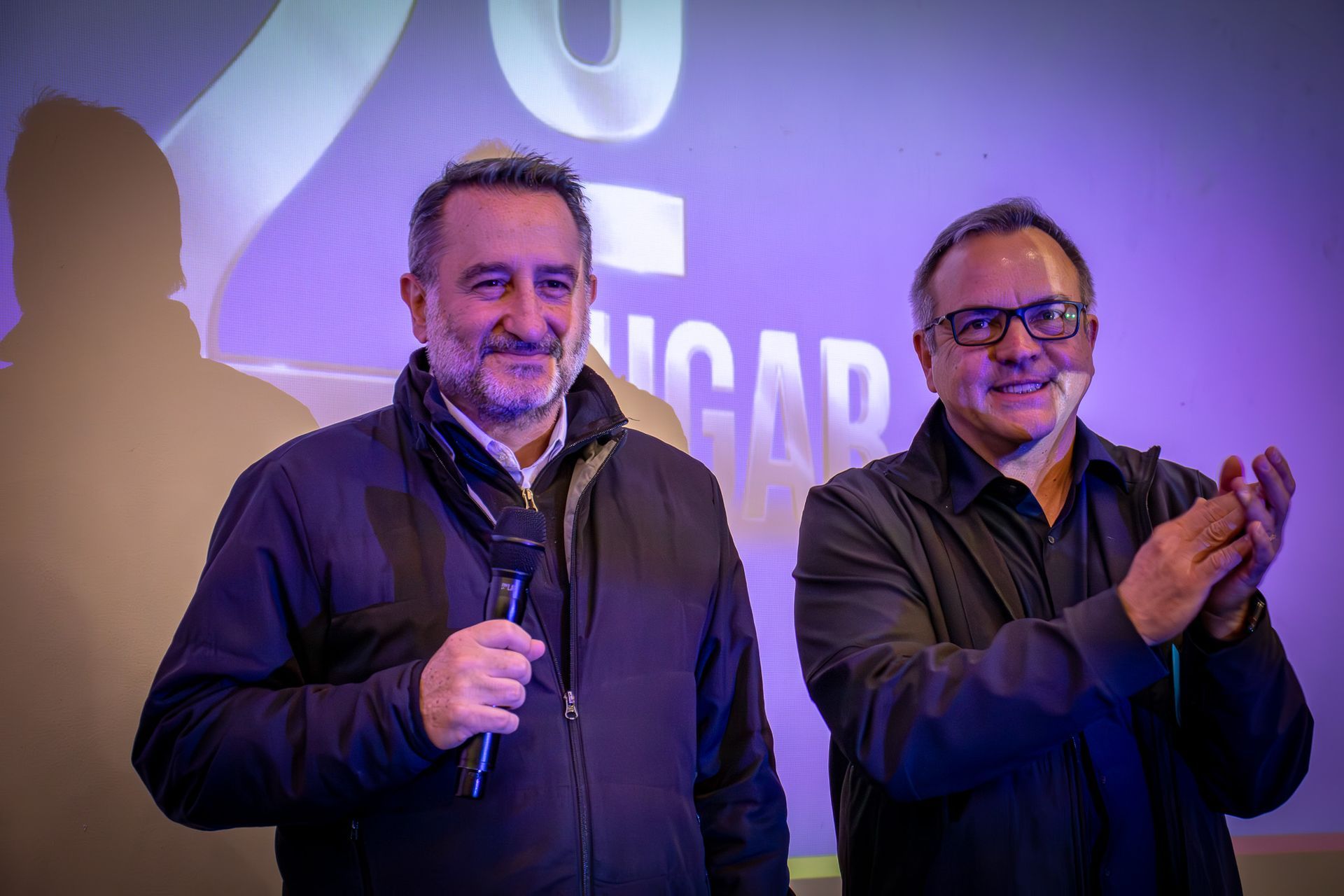 Dois homens em pé em um palco. Um segura um microfone e sorri, o outro bate palmas. Fundo roxo com texto.