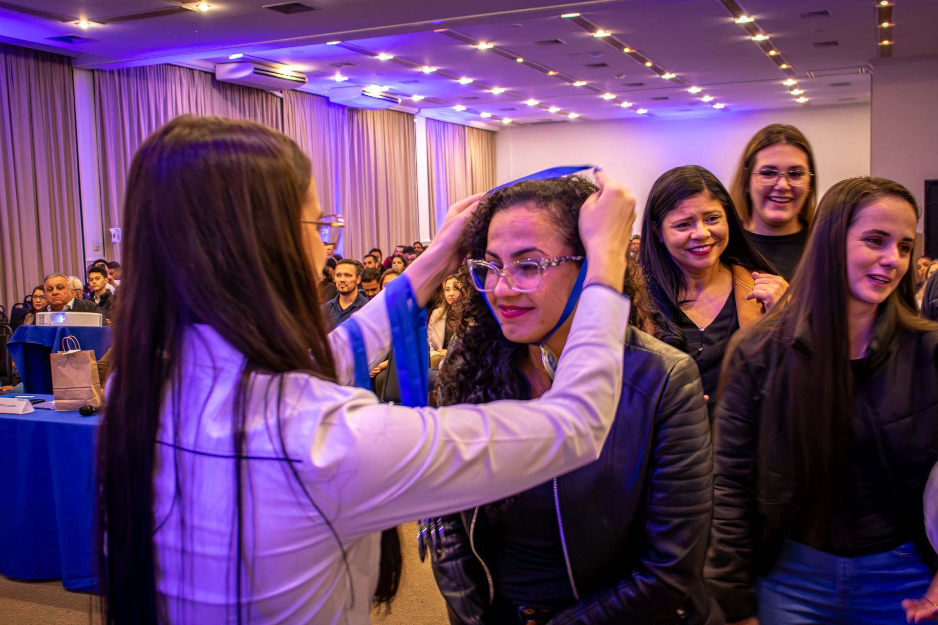 Uma mulher coloca um capelo de formatura em outra mulher durante uma cerimônia. Várias outras mulheres sorriem ao fundo, em um salão bem iluminado.