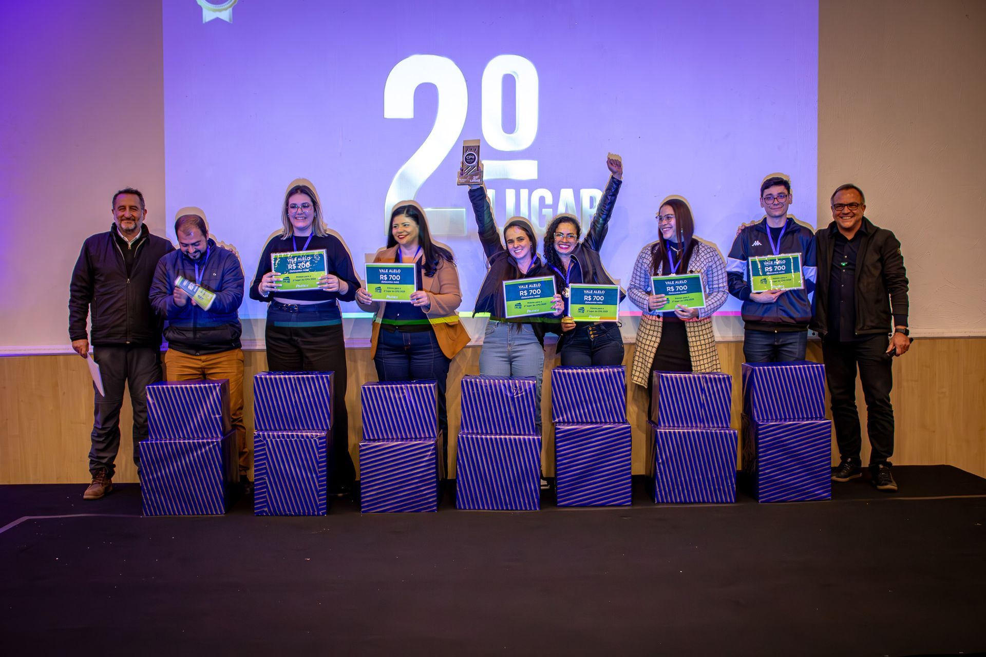 Um grupo de pessoas em um palco comemorando o segundo lugar. Elas seguram certificados e presentes em frente a um fundo azul.