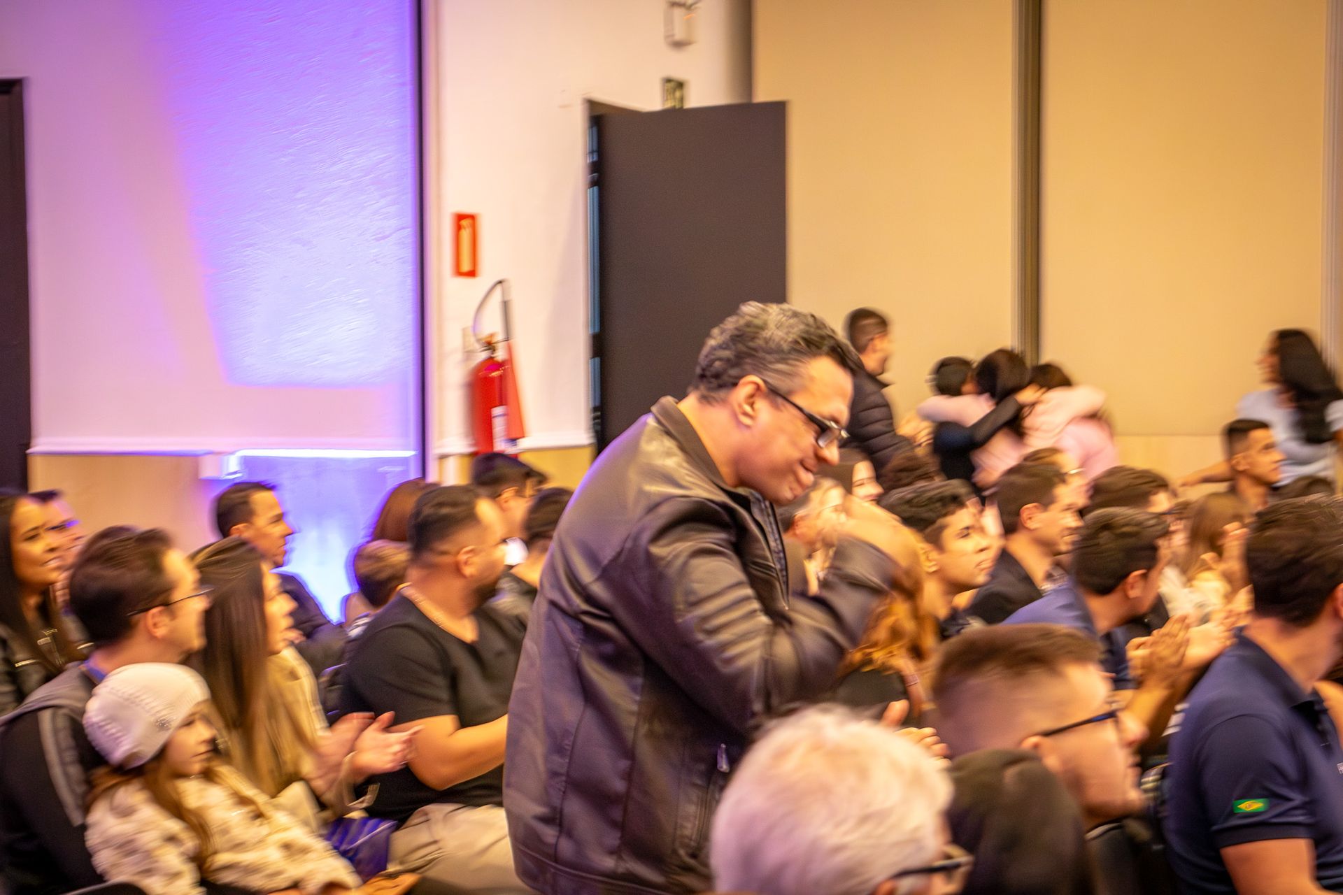 Homem de jaqueta de couro interagindo com uma multidão em um evento. Pessoas sentadas, algumas se abraçando, em um ambiente interno bem iluminado.