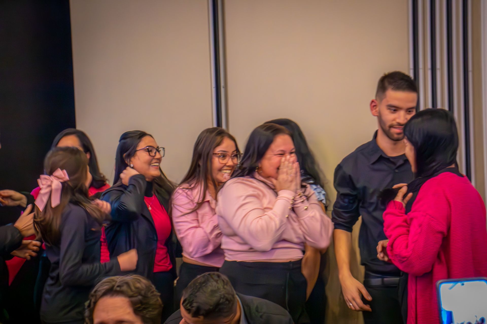 Uma mulher de camisa rosa chora, cercada por pessoas sorridentes. Uma celebração acontece em um ambiente fechado.