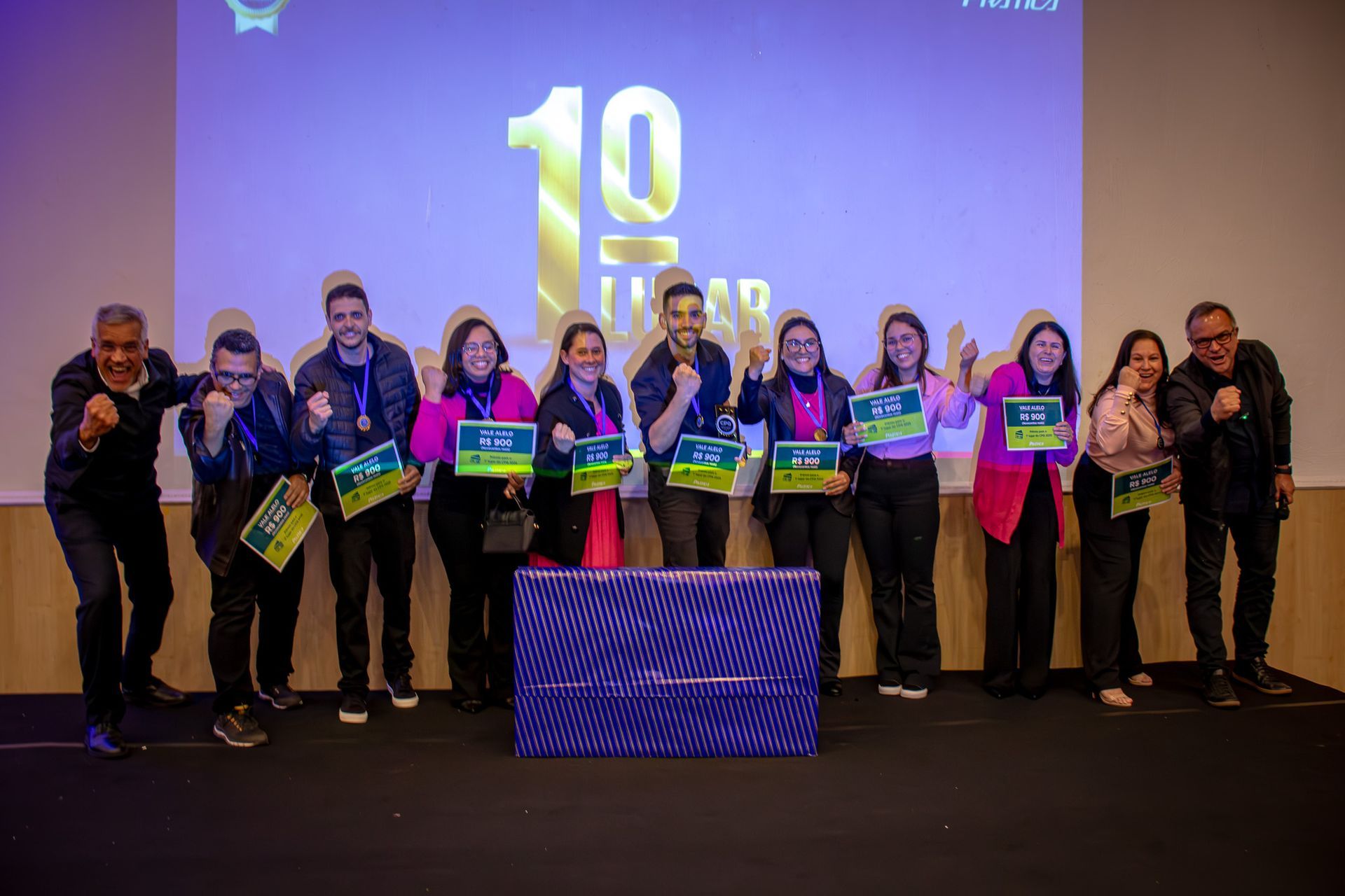 Grupo de pessoas comemorando 10 anos de casamento, segurando certificados e presentes. Elas estão em um palco com fundo azul.