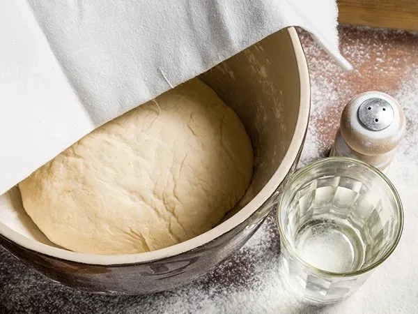 Uma tigela de massa coberta com uma toalha ao lado de um copo de água.