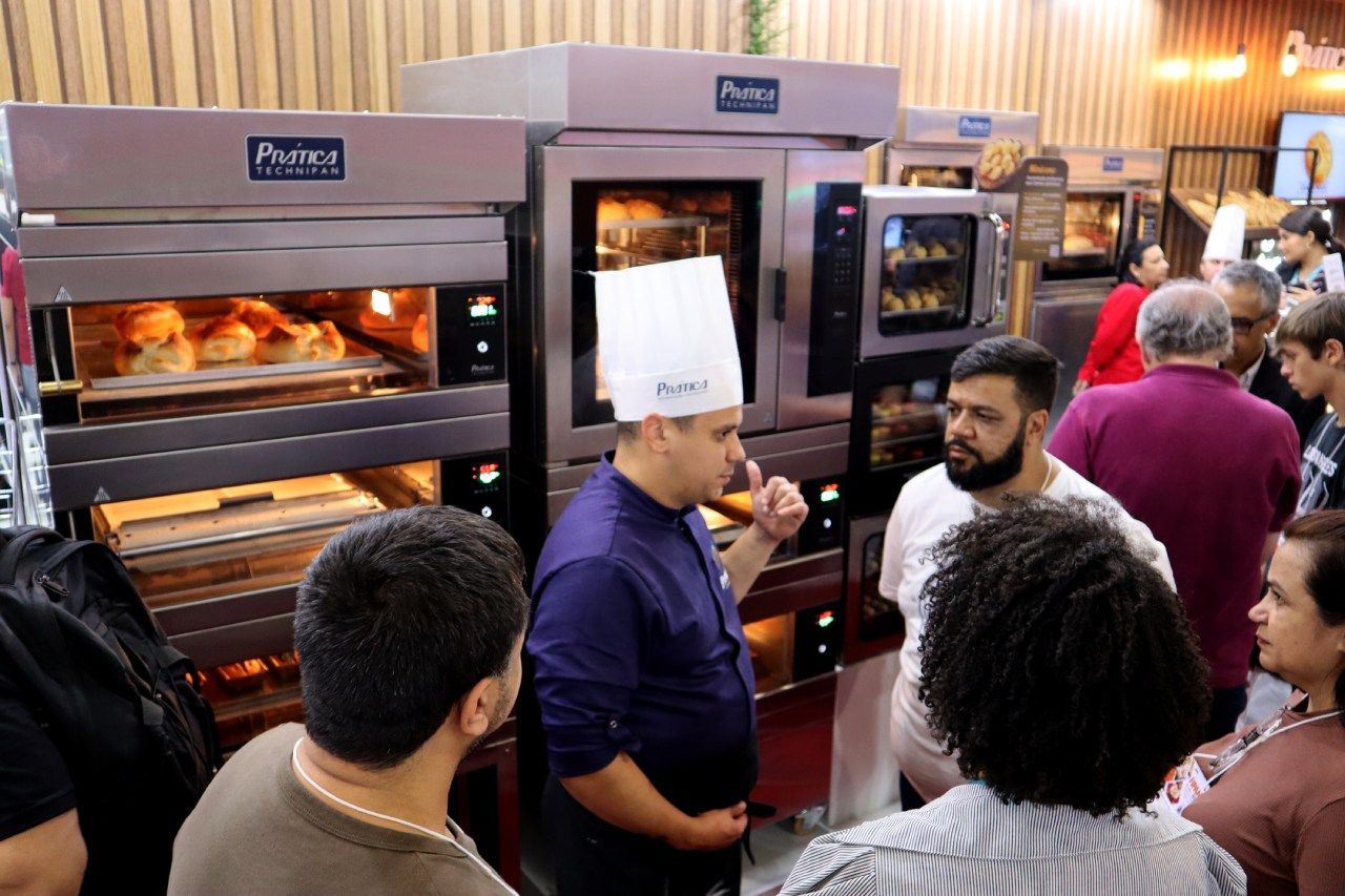 Um homem com chapéu de chef está conversando com um grupo de pessoas em frente a um forno.