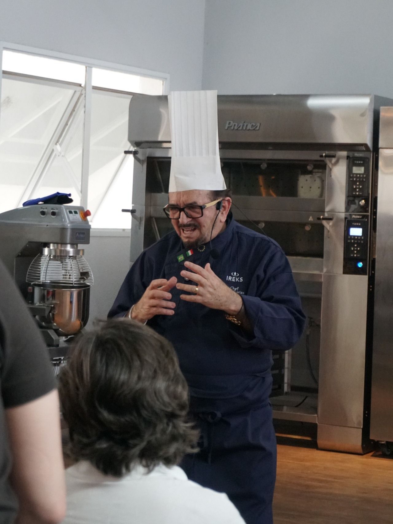 Chef de uniforme azul com chapéu alto, falando em uma cozinha, explicando um processo culinário.