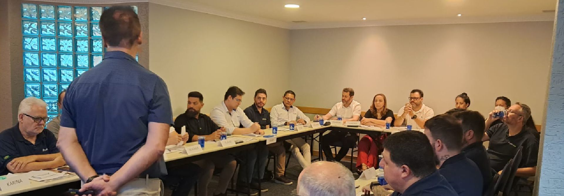 Um homem está de pé na frente de um grupo de pessoas sentadas em mesas em uma sala.