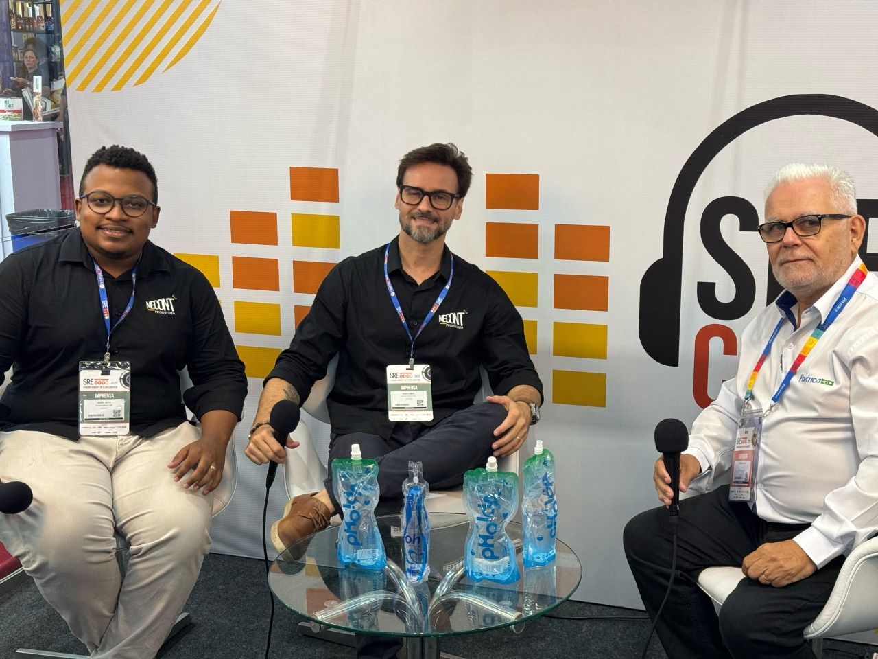 Três homens estão sentados ao redor de uma mesa com garrafas de água sobre ela.