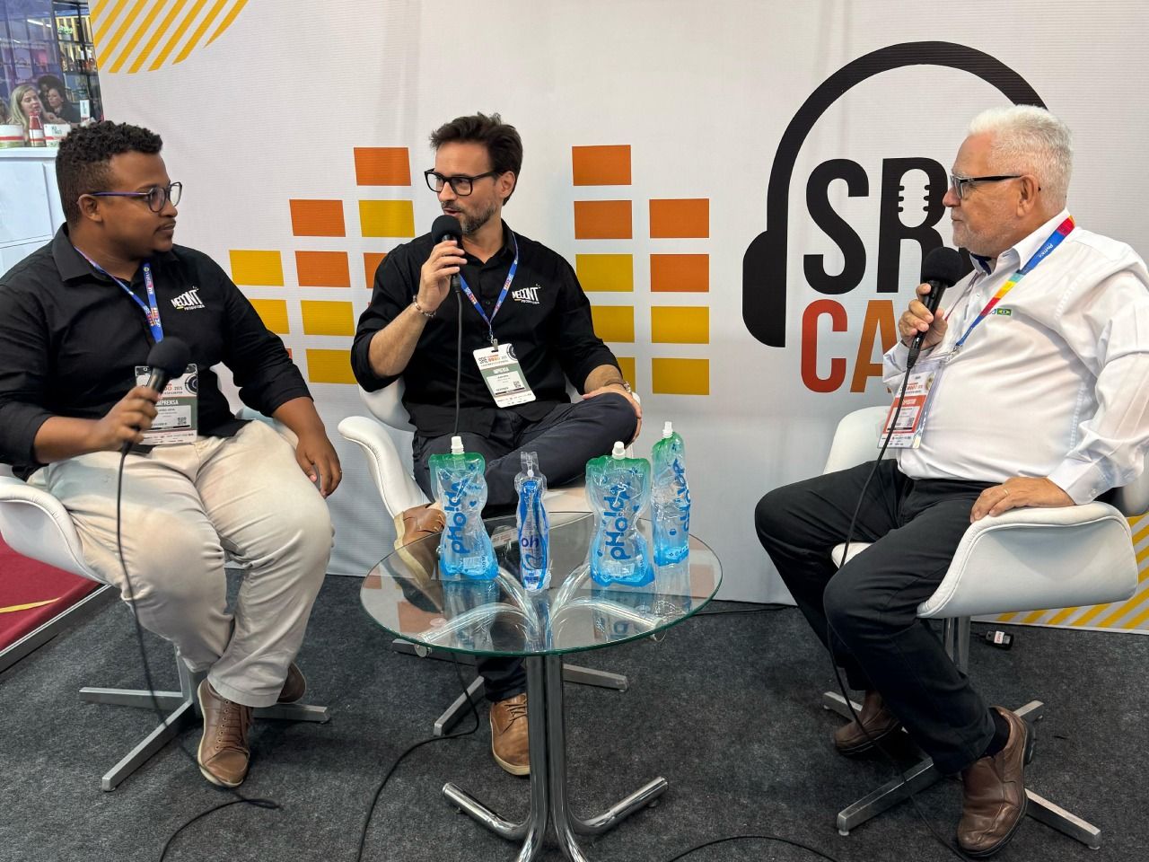 Três homens estão sentados em uma mesa com microfones em frente a uma placa que diz sr ca