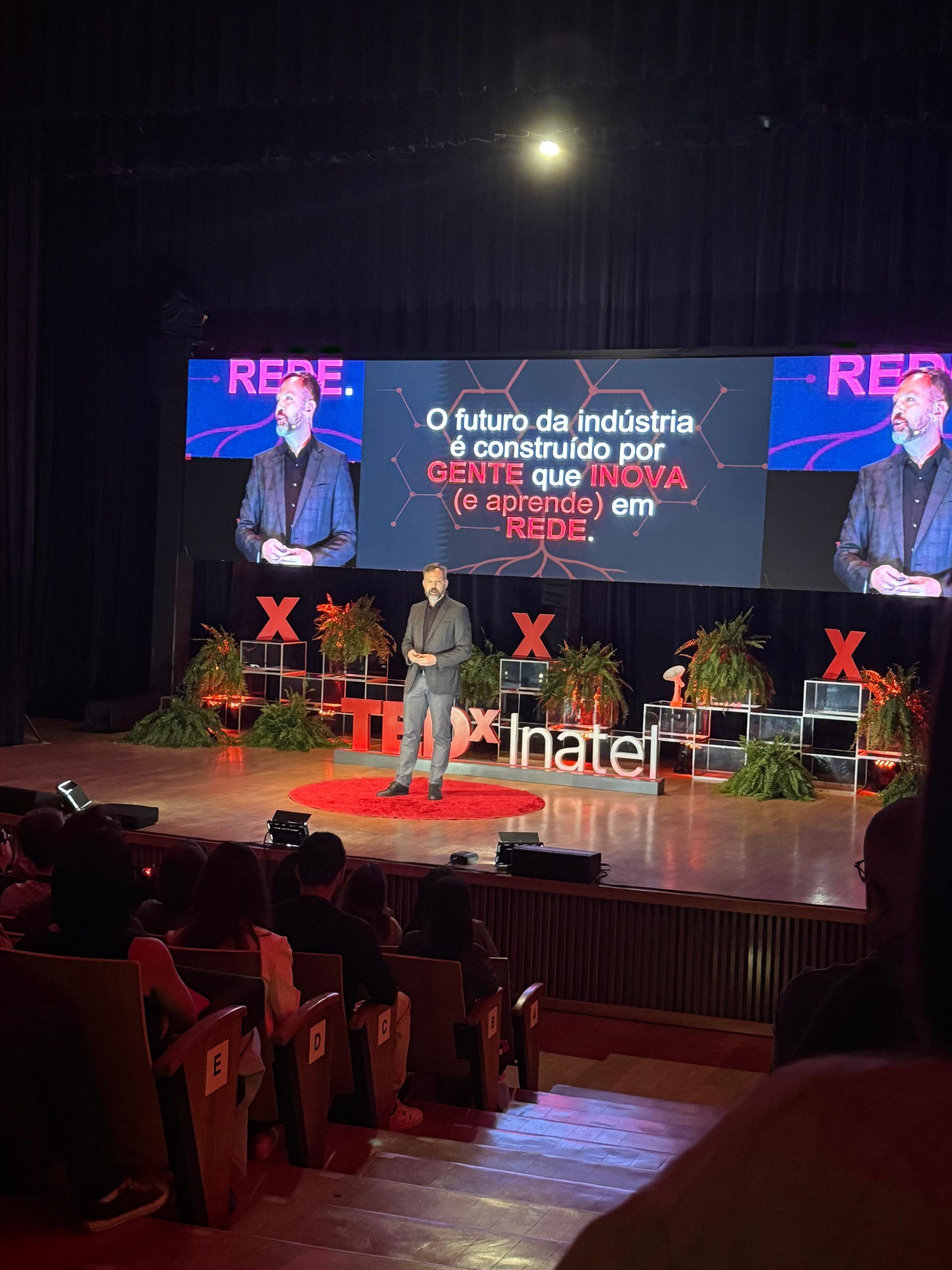 Homem discursando em um palco com um telão atrás. Público em primeiro plano.
