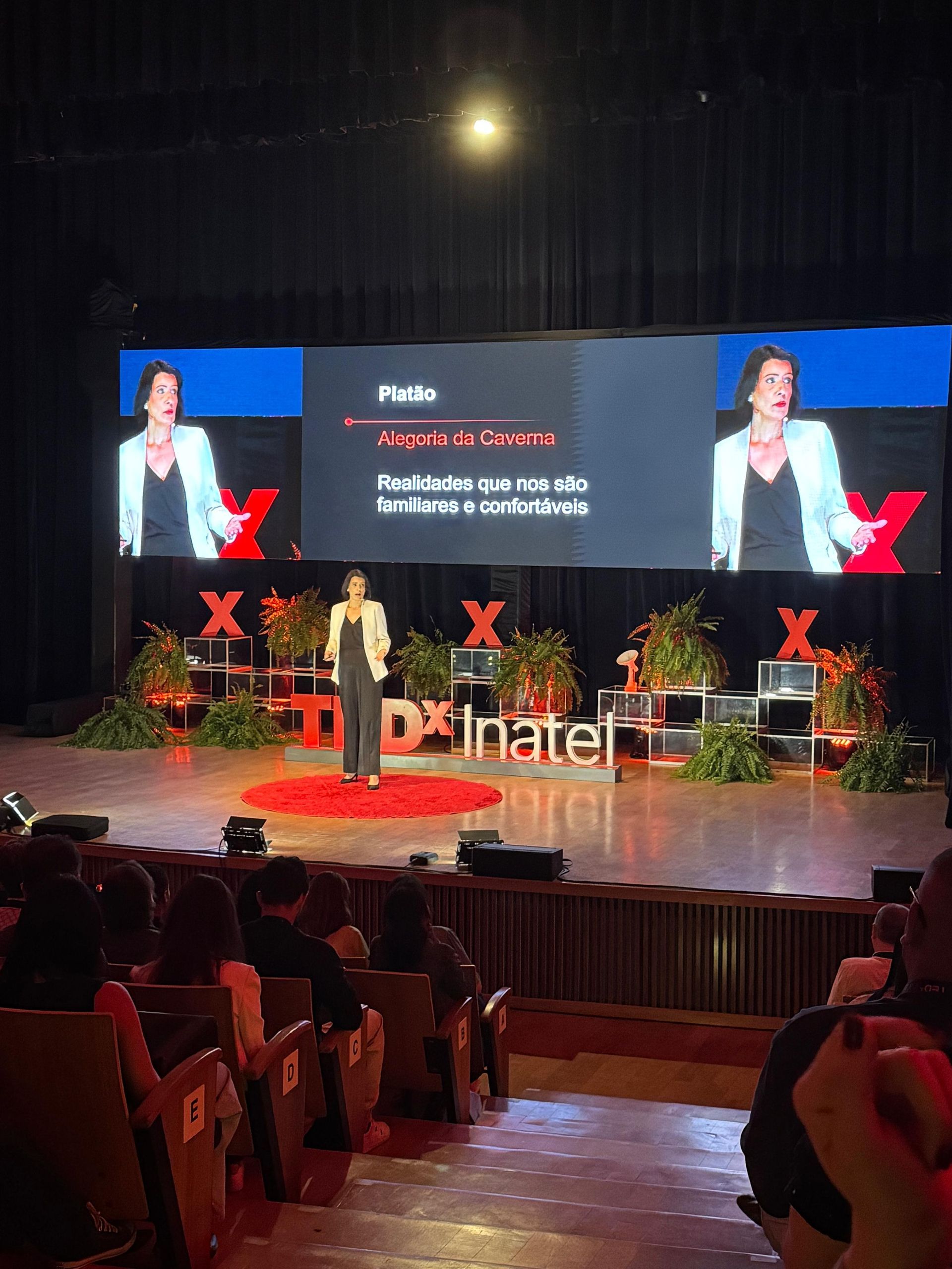 Mulher dando uma palestra no TEDx em um palco; público assistindo.