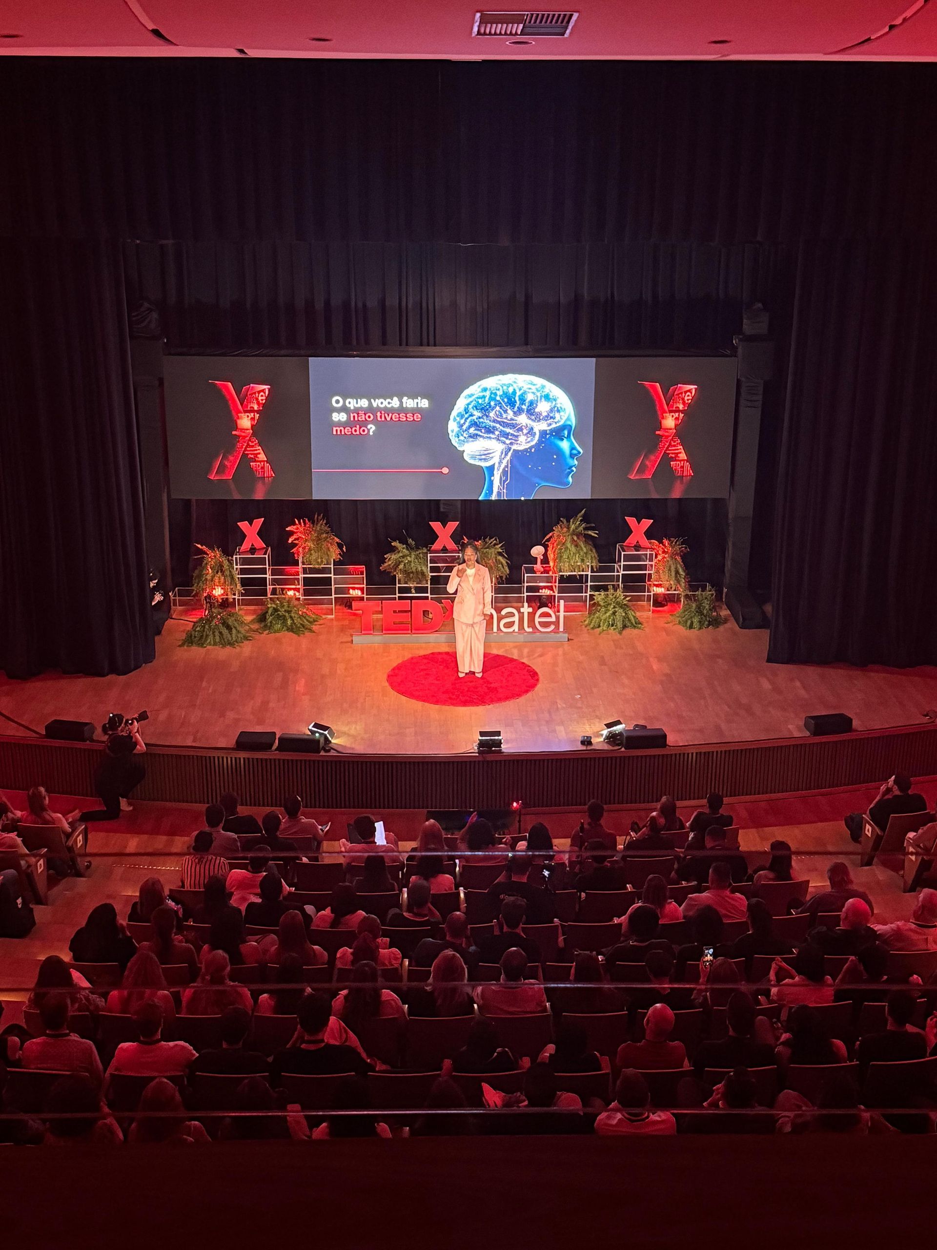 Uma pessoa falando em um palco em um evento TEDx. Público sentado, tela mostra a imagem de um cérebro.