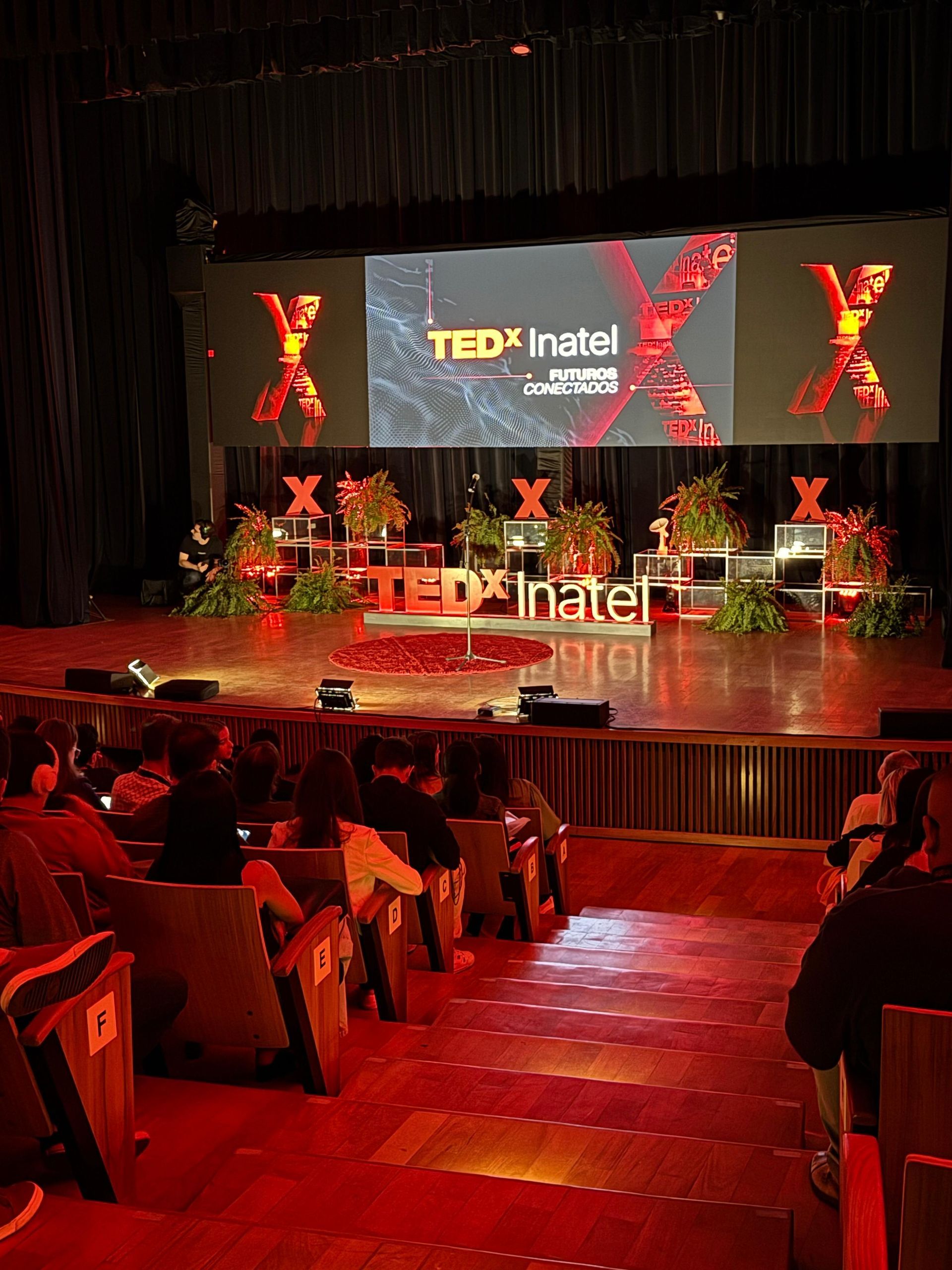 Palco do evento TEDx com iluminação vermelha e plateia, com logotipo e decoração floral.