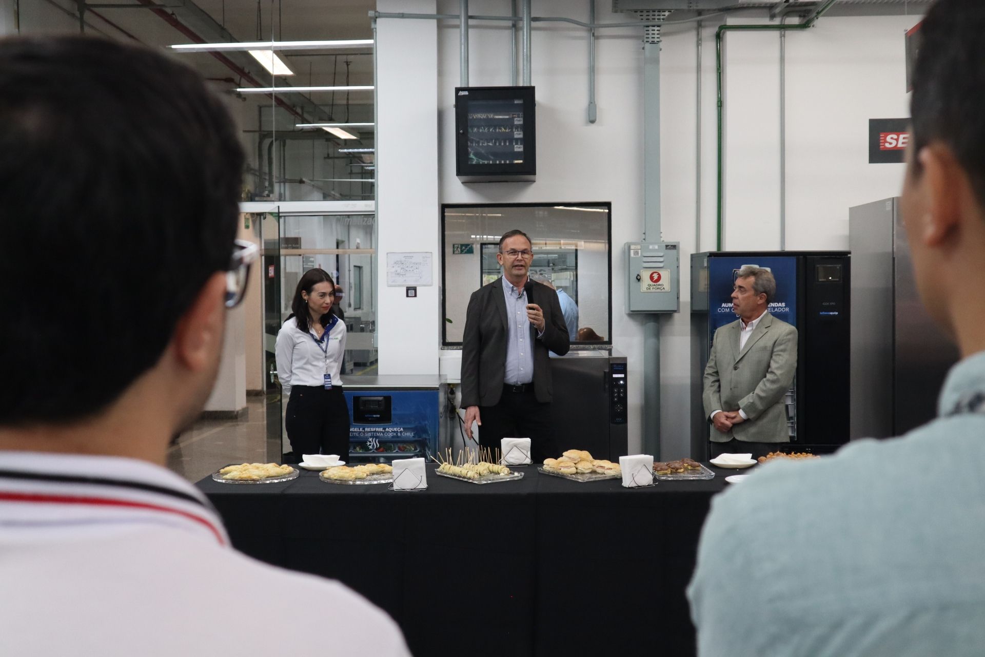 Pessoas em um evento; um homem falando em uma mesa com lanches, com outras duas pessoas ao lado. Ambiente industrial.