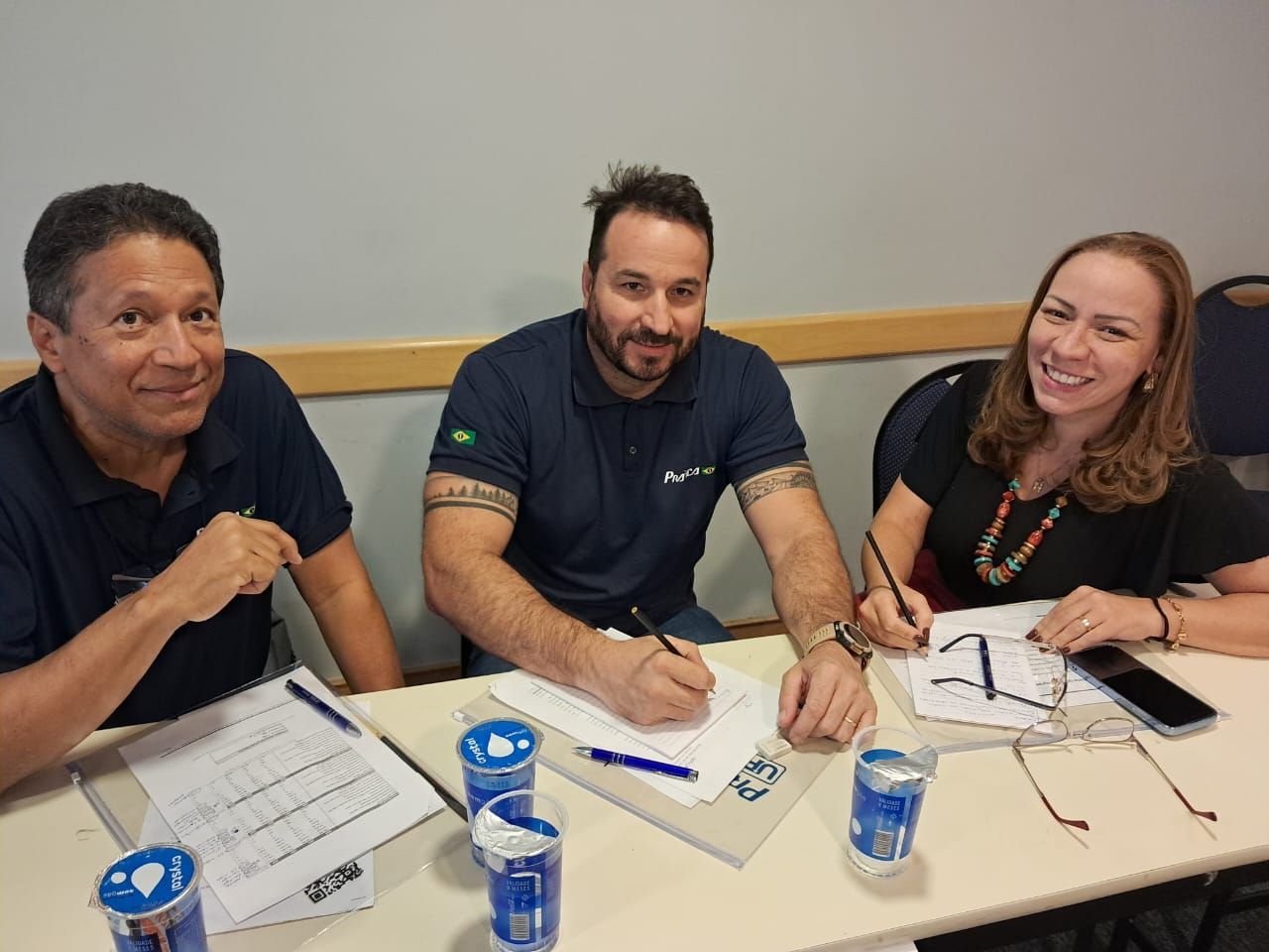 Três pessoas estão sentadas à mesa escrevendo em papéis.