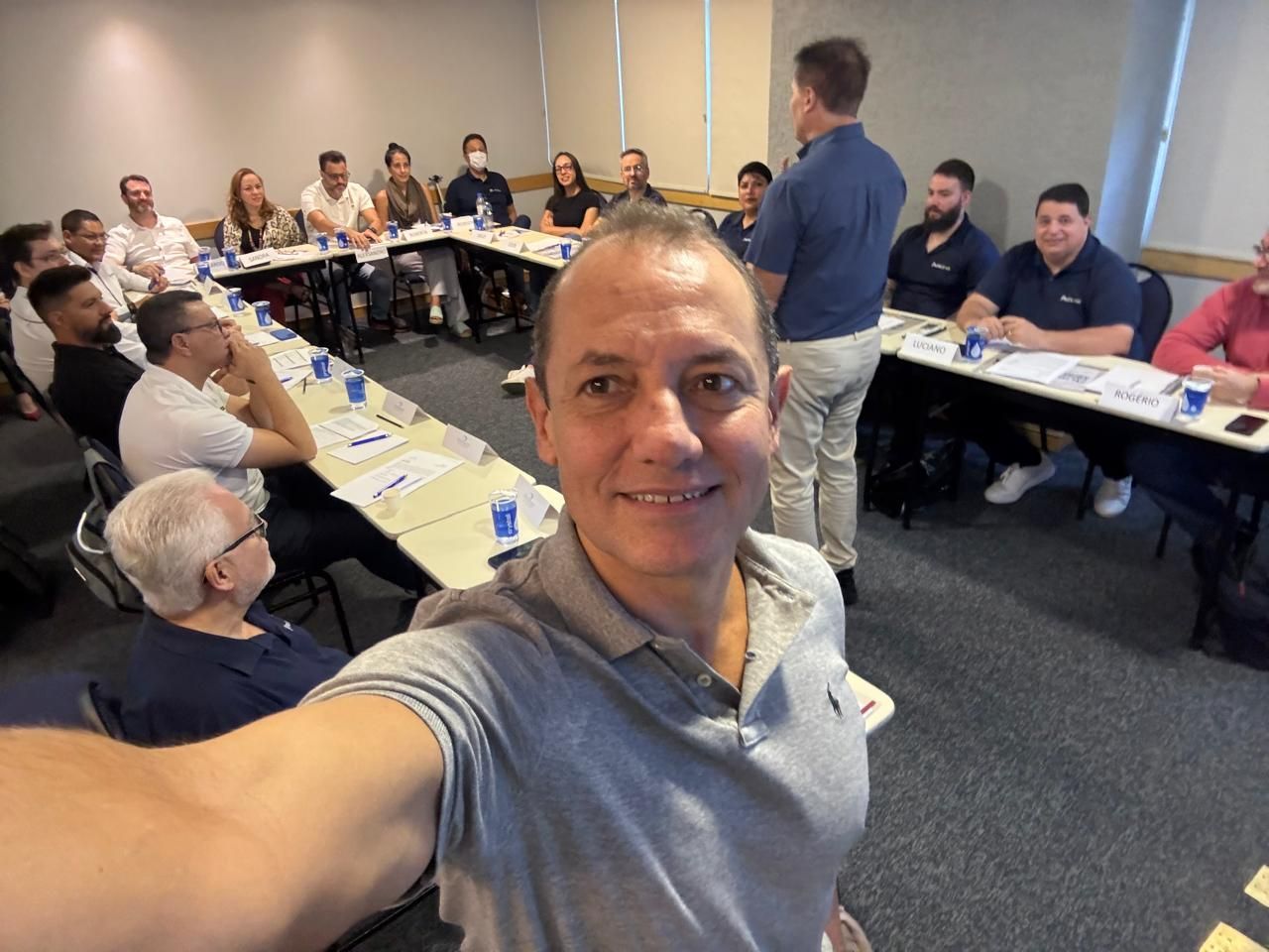 Um homem está tirando uma selfie na frente de um grupo de pessoas sentadas em mesas.