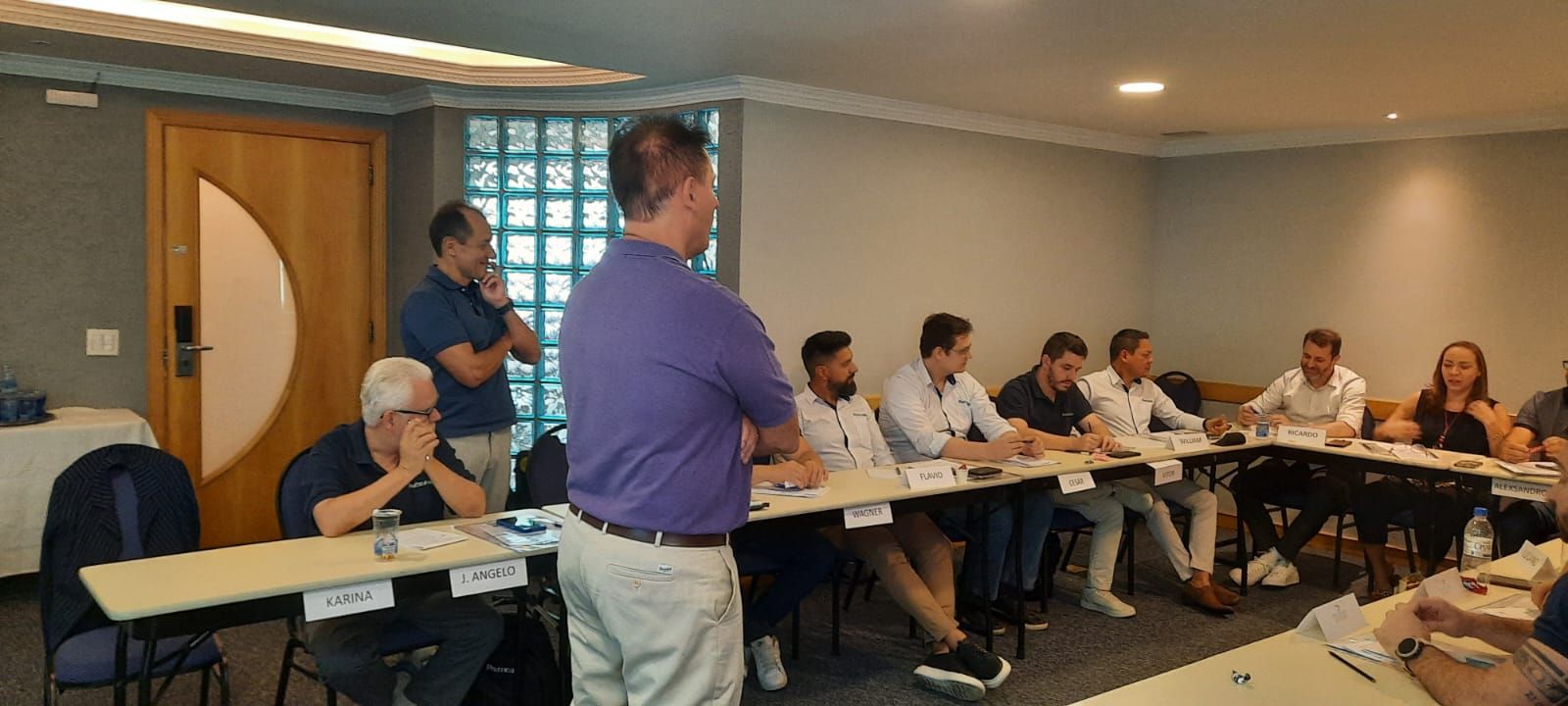 Um homem está de pé na frente de um grupo de pessoas sentadas em mesas em uma sala.