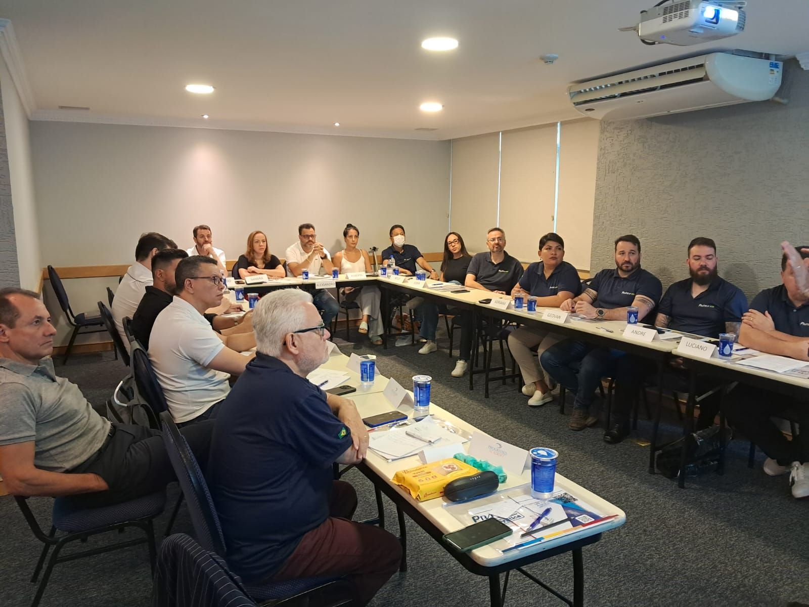 Um grupo de pessoas está sentado em longas mesas em uma sala de conferências.