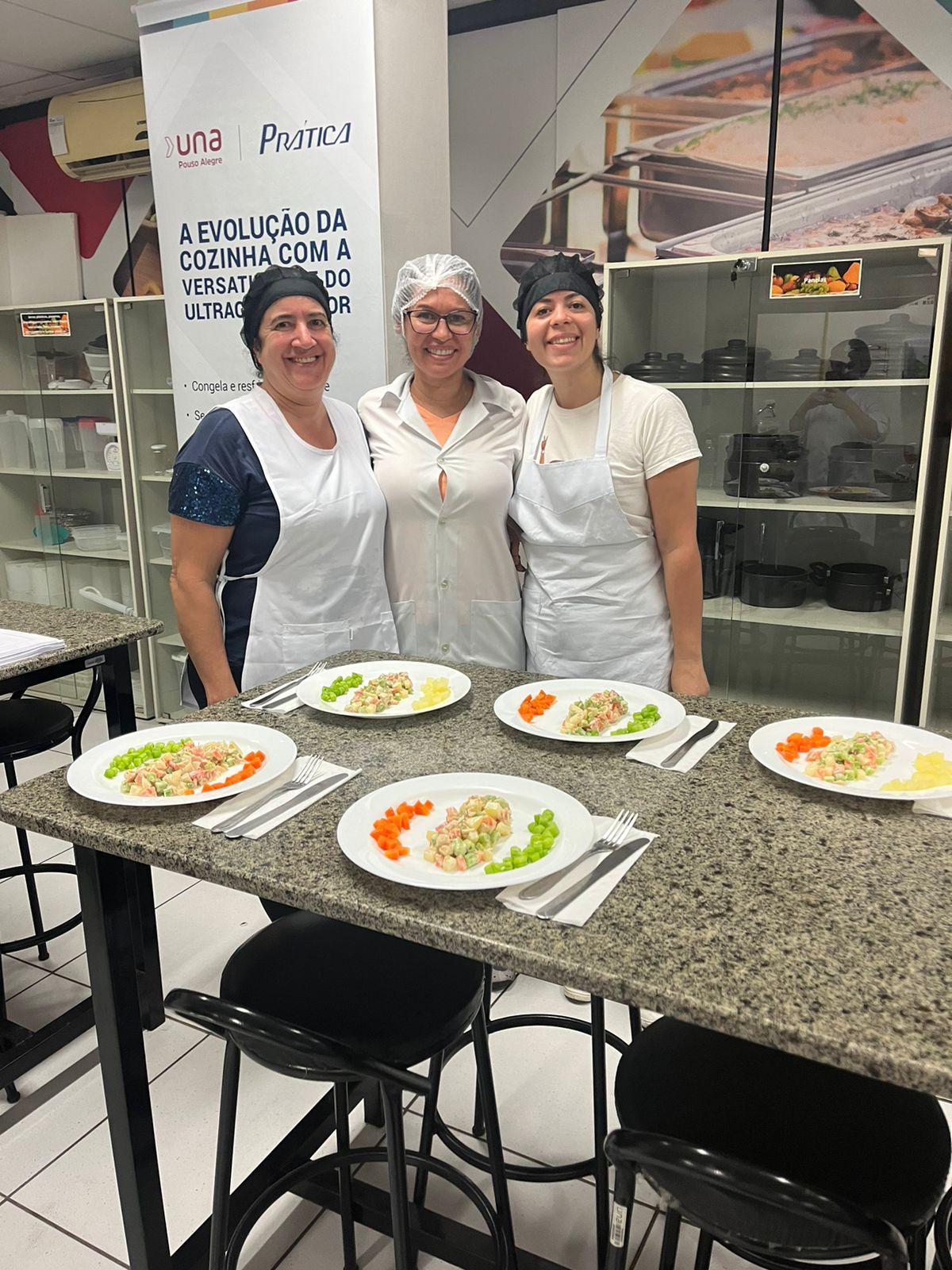 Três mulheres estão posando para uma foto em frente a uma mesa com pratos de comida.