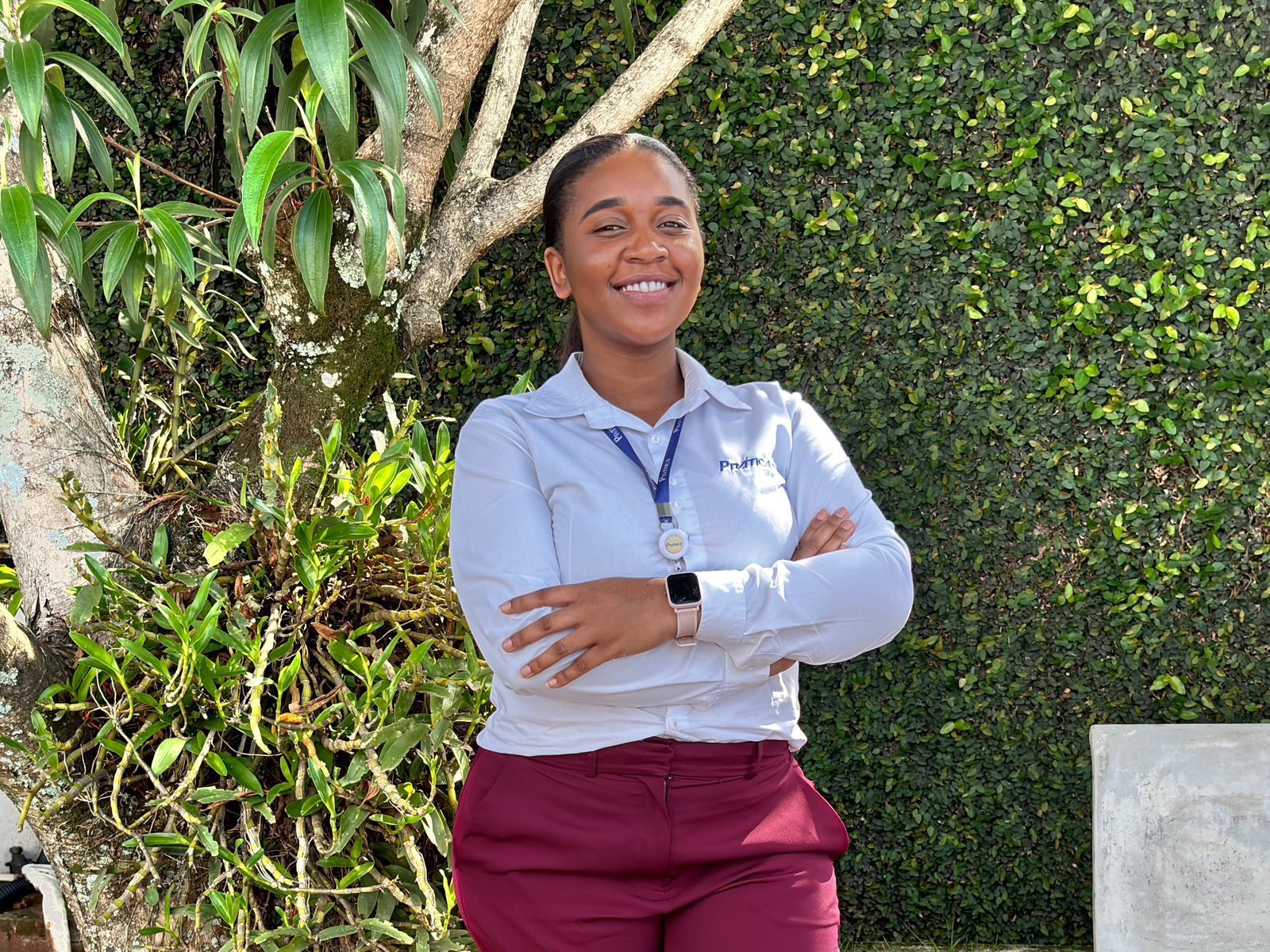 Uma mulher está parada em frente a uma cerca viva com os braços cruzados e sorrindo.