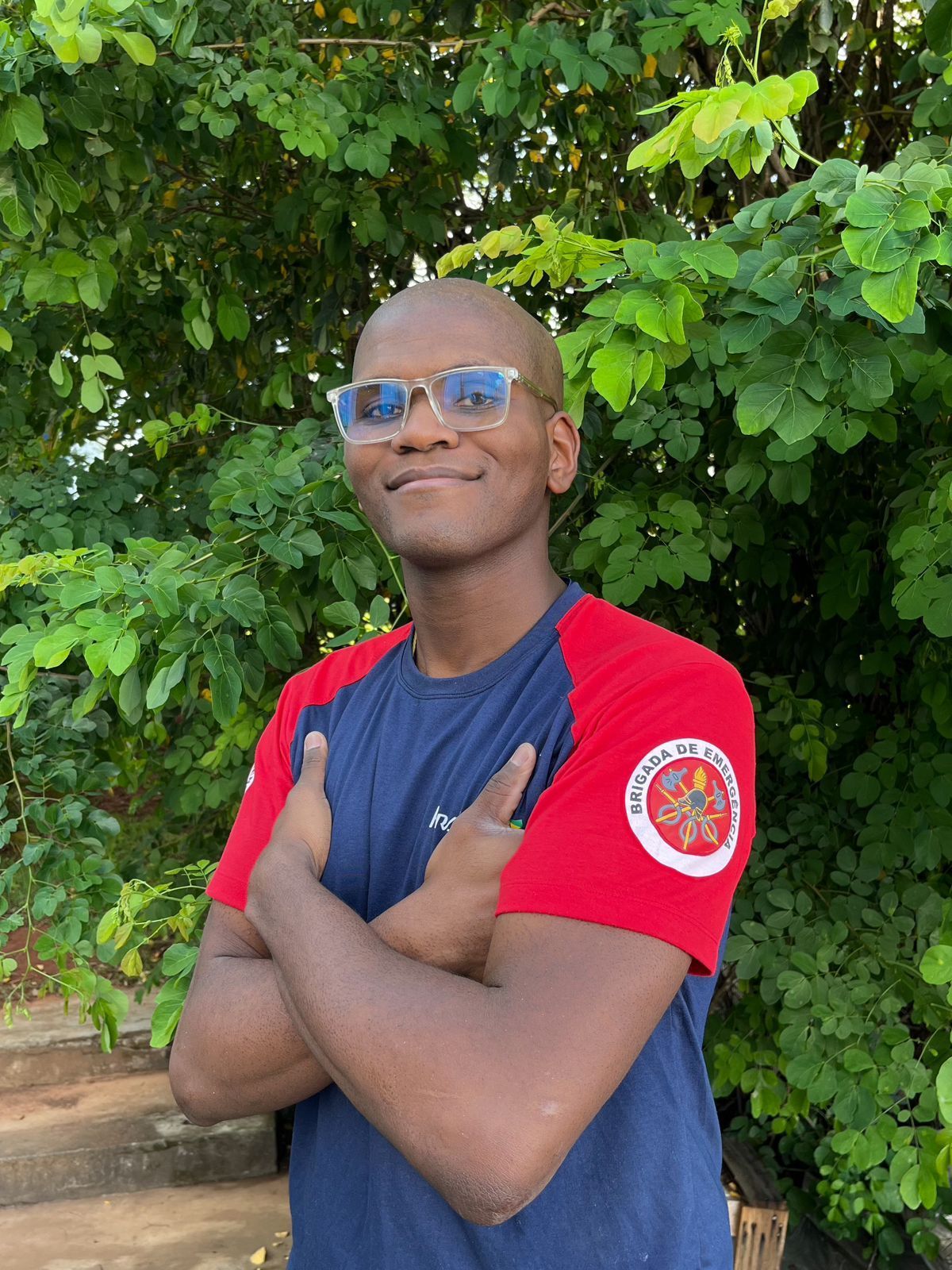 Um homem usando óculos escuros e uma camisa azul e vermelha está de pé com os braços cruzados em frente a uma árvore.