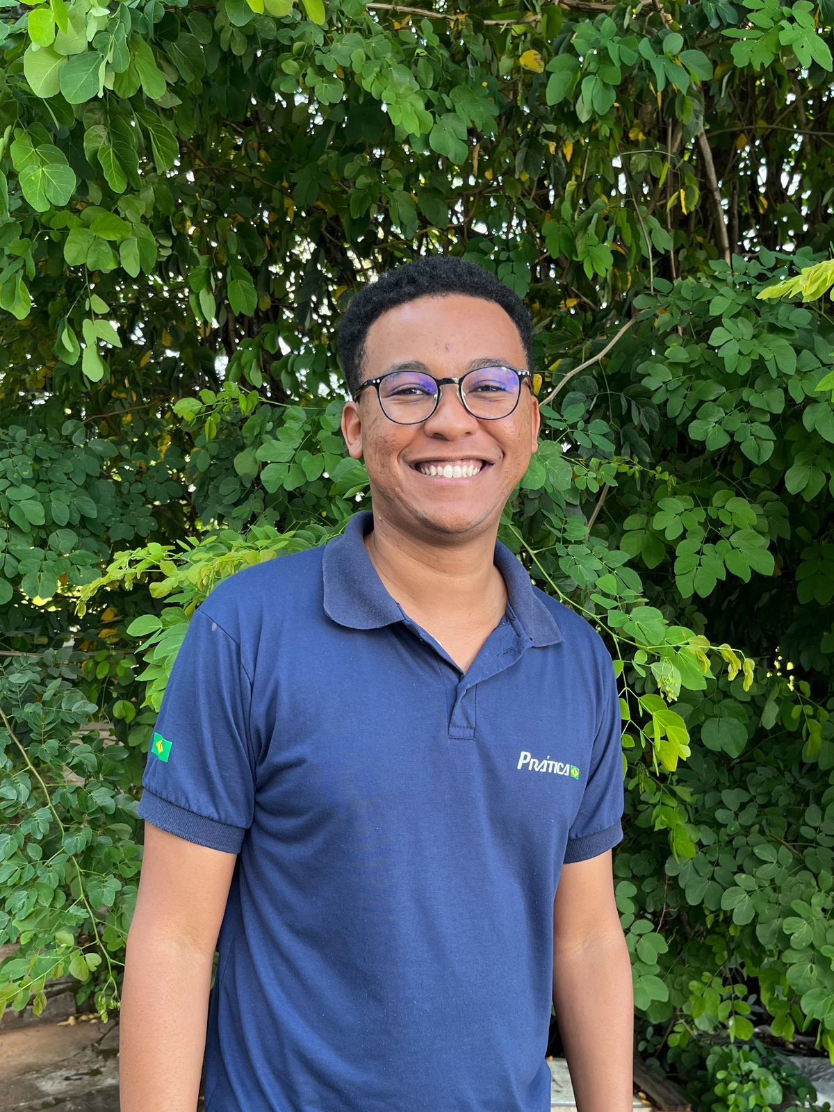 Um jovem usando óculos e uma camisa azul está sorrindo em frente a uma árvore.
