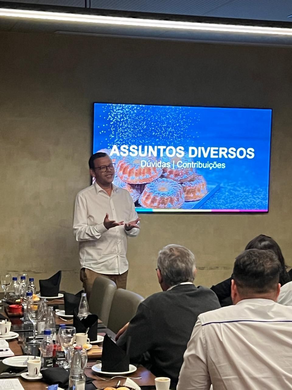 Um homem está fazendo uma apresentação em frente a uma tela que diz assuntos diversos