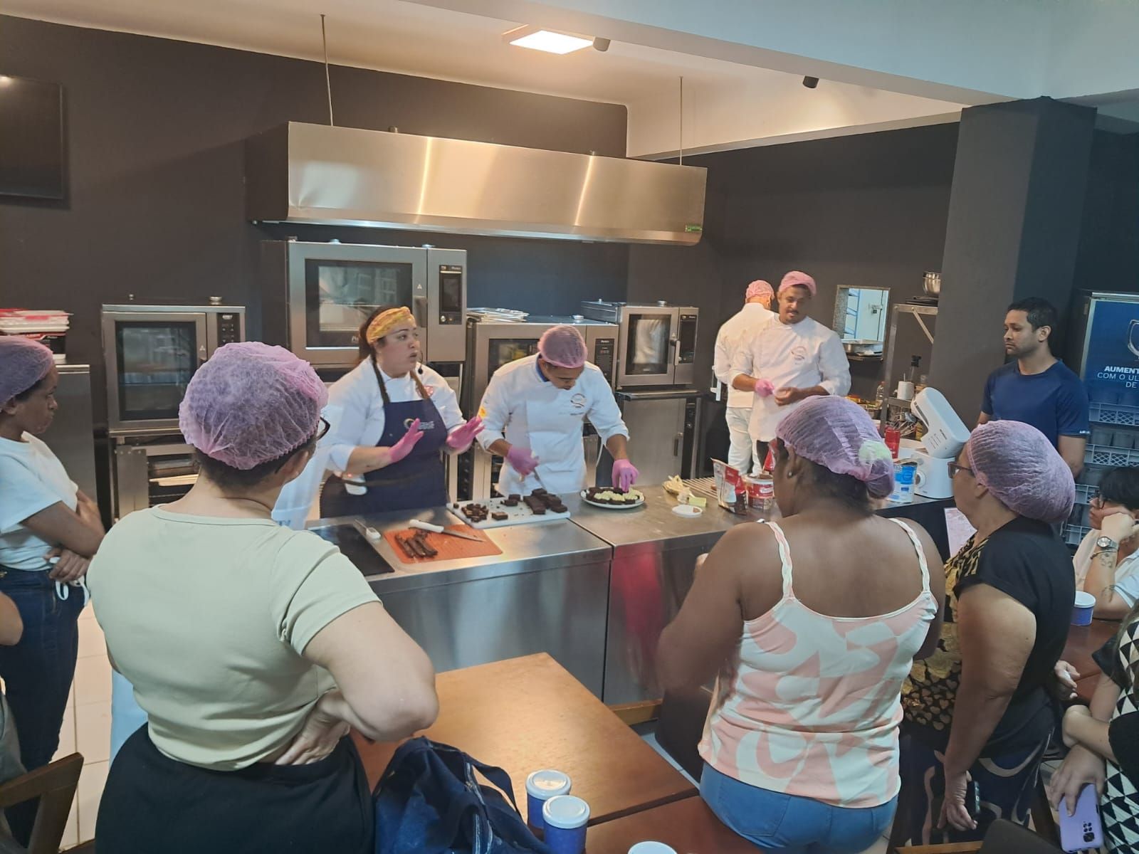 Pessoas em uma cozinha comercial observam chefs preparando comida. Alguns participantes usam toucas cor-de-rosa.