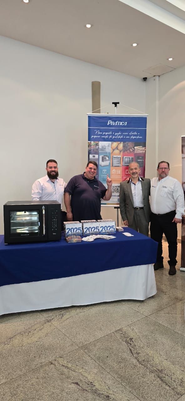 Quatro homens estão atrás de uma mesa com um pequeno forno, uma faixa e materiais promocionais em uma sala bem iluminada.