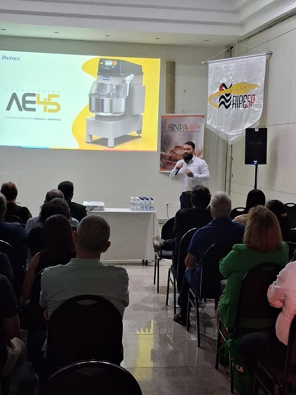 Homem fazendo uma apresentação para uma plateia em uma conferência, uma tela exibe o produto AE45.