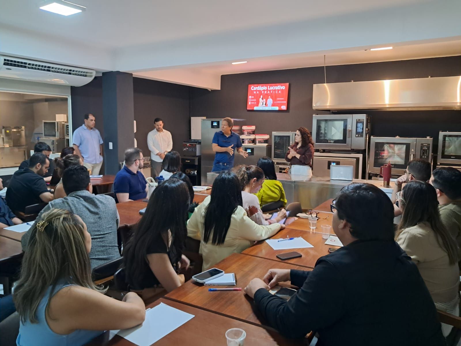 Um grupo de pessoas em uma reunião. Os apresentadores falam enquanto os participantes ouvem e tomam notas.