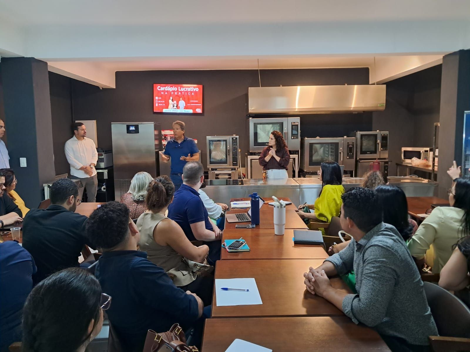 Pessoas em reunião, algumas falando. Ambiente escuro com equipamentos de cozinha ao fundo.