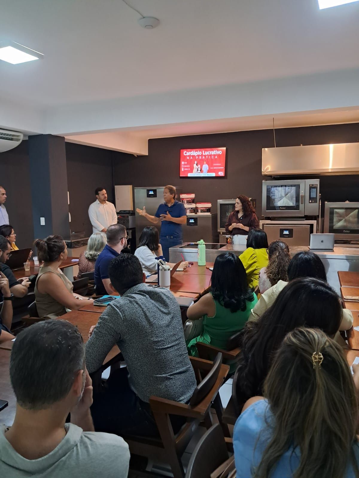 Um grupo de pessoas em uma reunião. Uma mulher fala à frente. Uma tela de TV é visível.