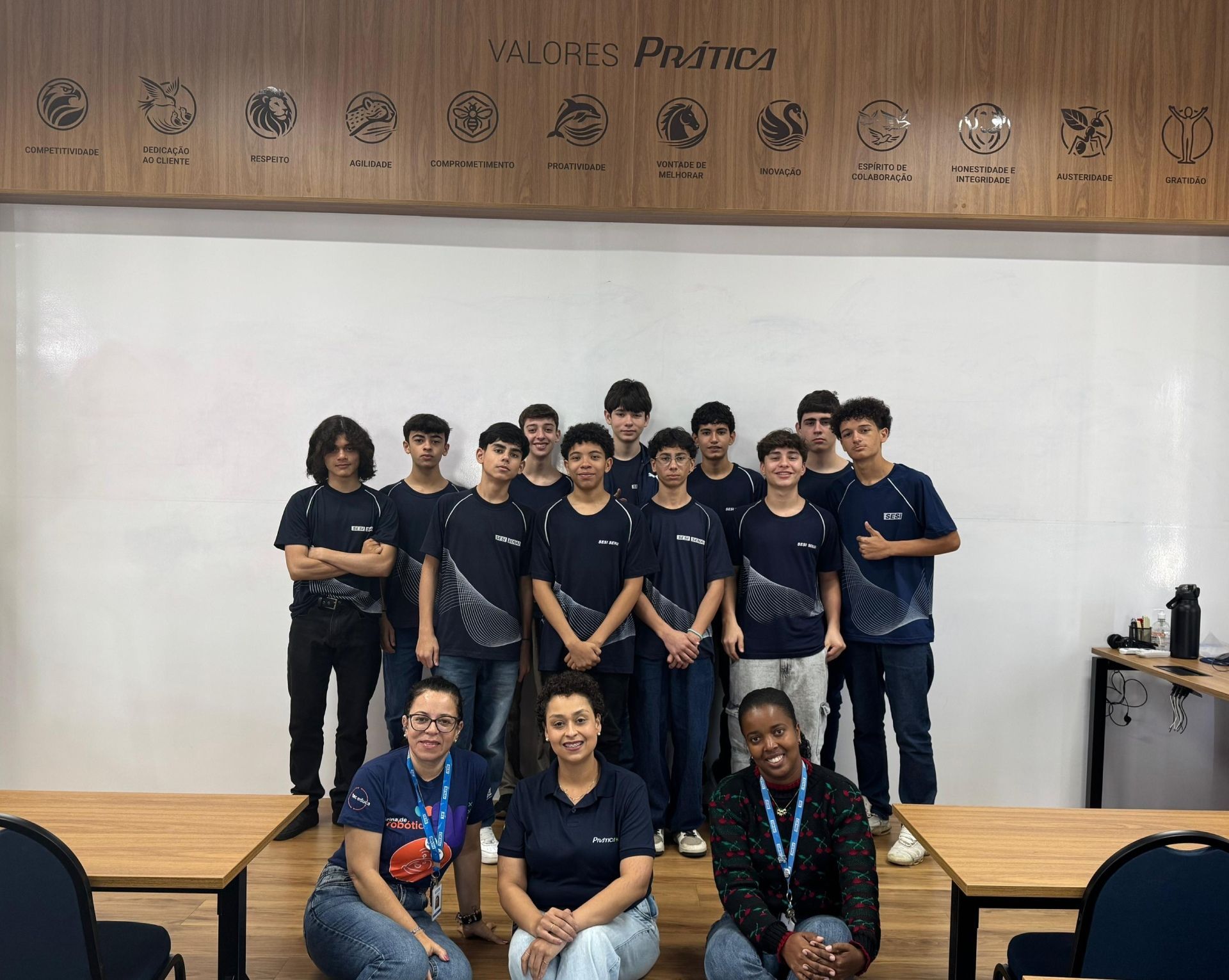 Um grupo de adolescentes e três mulheres posam juntos em um ambiente fechado; os adolescentes usam camisetas combinando.