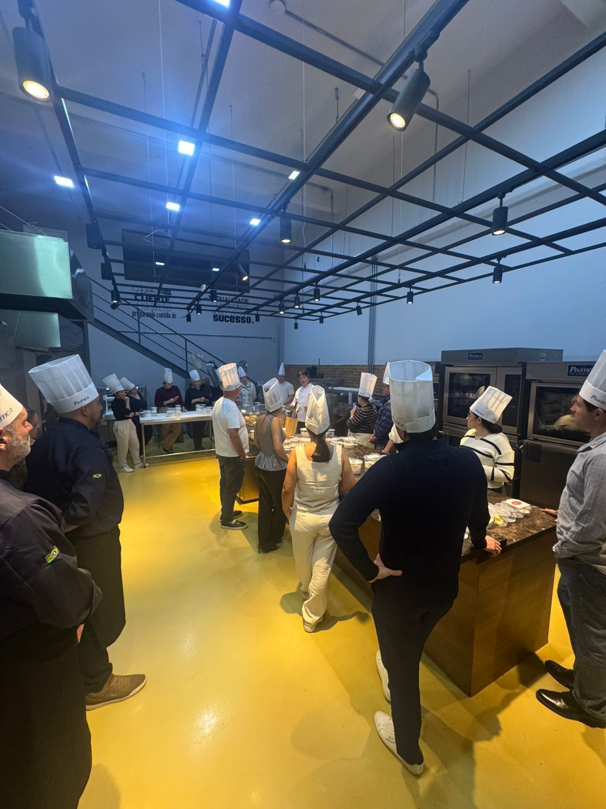 Chefs de chapéu branco em uma aula de culinária, reunidos ao redor de um balcão. Piso amarelo, ambiente em estilo industrial.