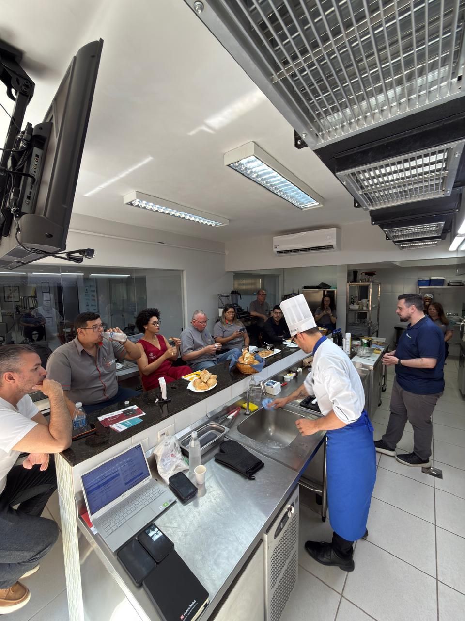 Um chef prepara comida para um grupo em uma cozinha. As pessoas observam em um bar, conversando, em um espaço bem iluminado.