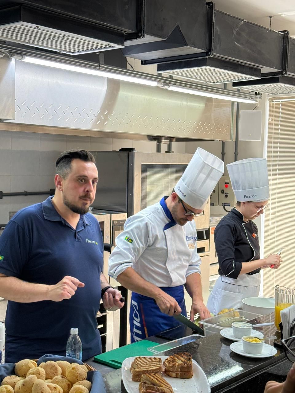 Três chefs em uma cozinha; um cortando a comida, o outro observando. Uma variedade de alimentos, panelas e fornos são visíveis.