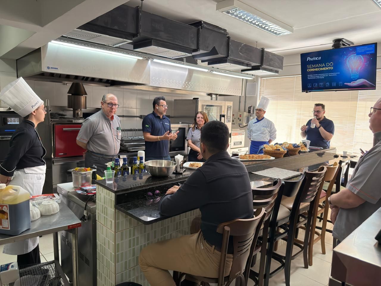 Pessoas em uma cozinha profissional; um homem fala, outros ouvem. Chefs, comida e eletrodomésticos são visíveis.