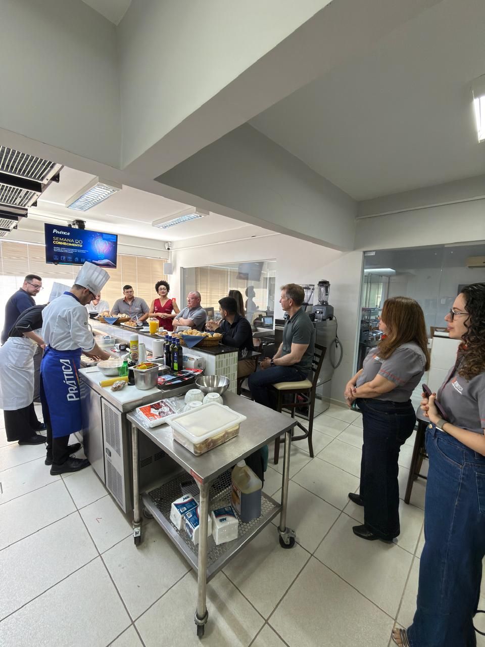 Chef demonstra culinária em uma cozinha. Pessoas observam. Iluminação forte, ingredientes variados.