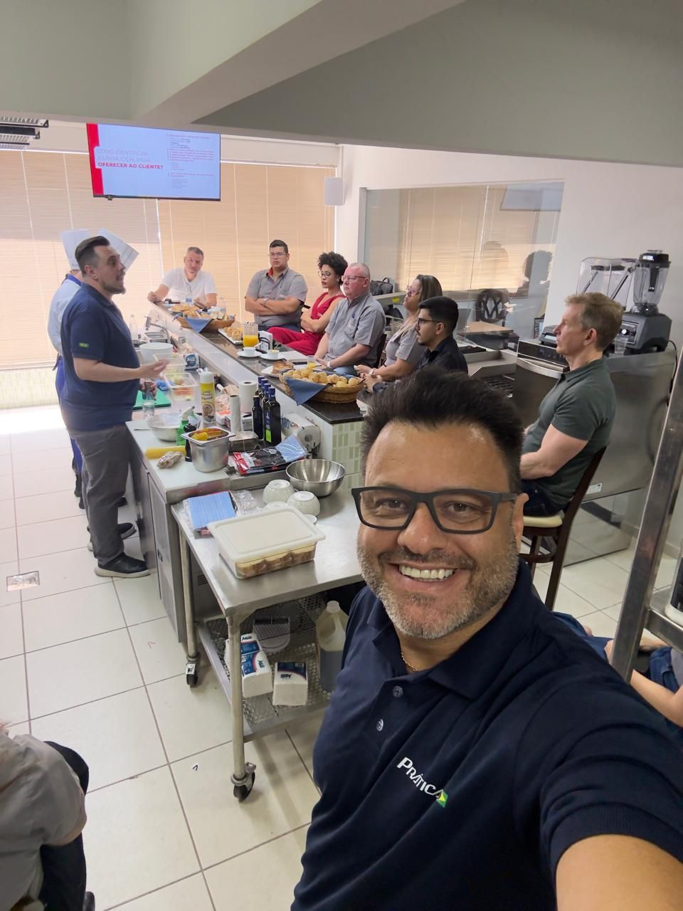 Homem sorrindo, tirando uma selfie na cozinha; uma aula de culinária em andamento, pessoas reunidas ao redor de uma mesa de aço inoxidável.